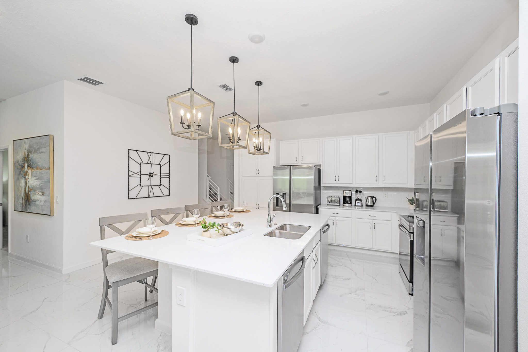 A well-appointed white kitchen featuring a large central island with a sink and seating, surrounded by white shaker cabinets and stainless steel appliances, with modern pendant lights overhead.