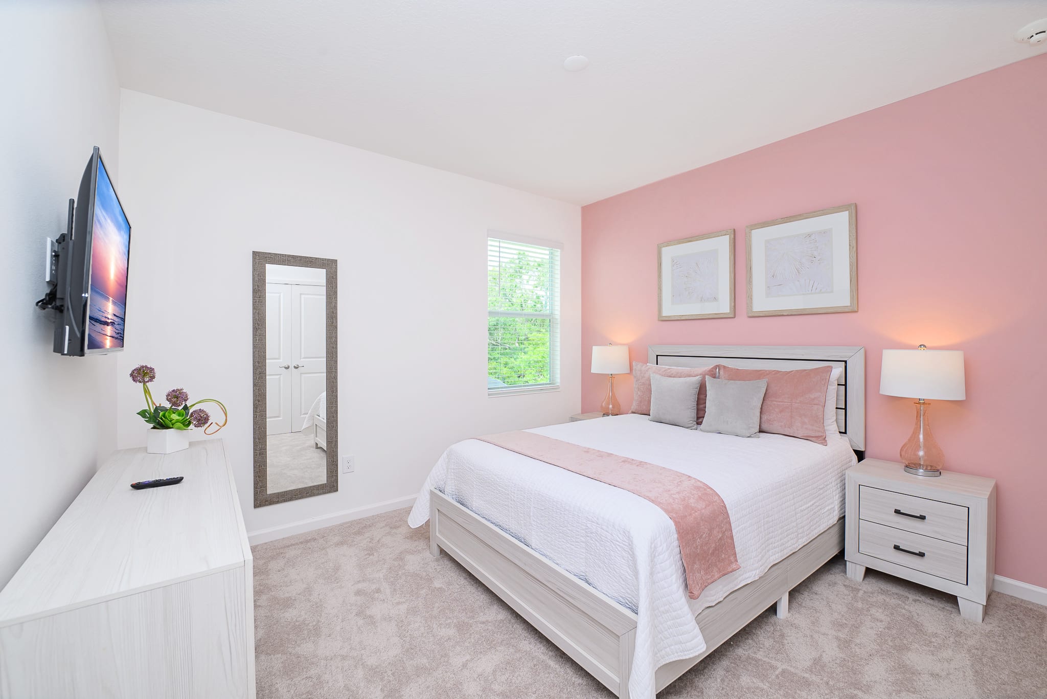 Inviting bedroom with a pink accent wall behind a light gray bed with pink and white bedding. Matching nightstands with lamps flank the bed, and a window with blinds offers natural light.