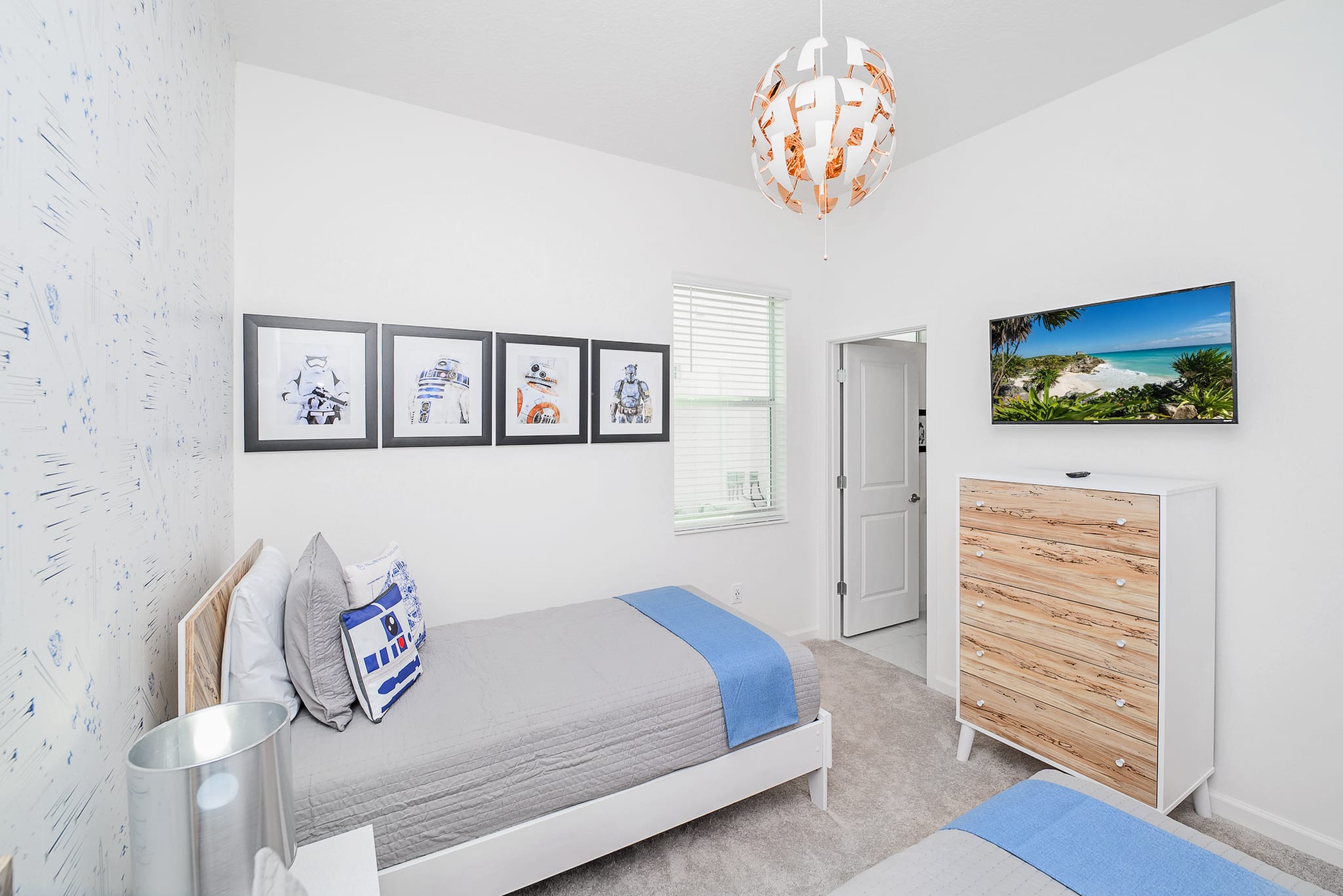 Themed kids' bedroom with two twin beds, gray bedding, and blue accents. Star Wars-themed artwork adorns the wall, and a unique white and orange pendant light hangs from the ceiling.