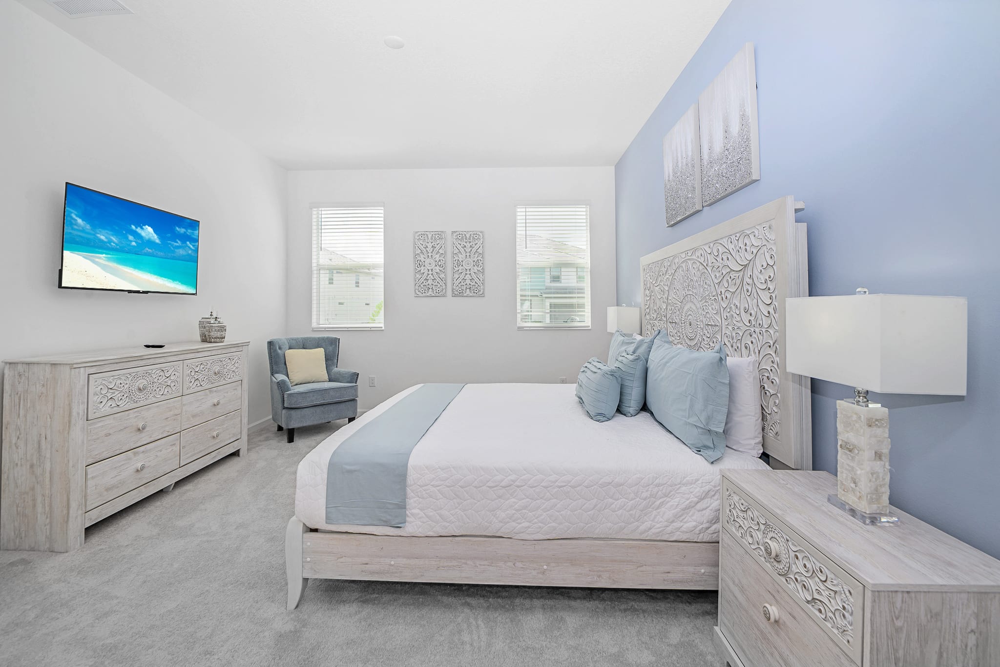 Bright bedroom with a white bed, ornate headboard, and light blue bedding. A wooden dresser with a wall-mounted TV is opposite the bed, and a light blue armchair sits in the corner by the windows.
