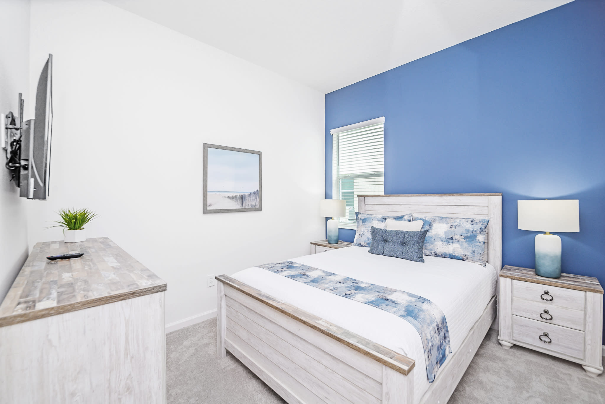 Bright bedroom with a white wooden bed frame, a striking blue accent wall, and a wall-mounted TV above a rustic white dresser. The bedding features blue and white patterns, complemented by a framed beach art piece.