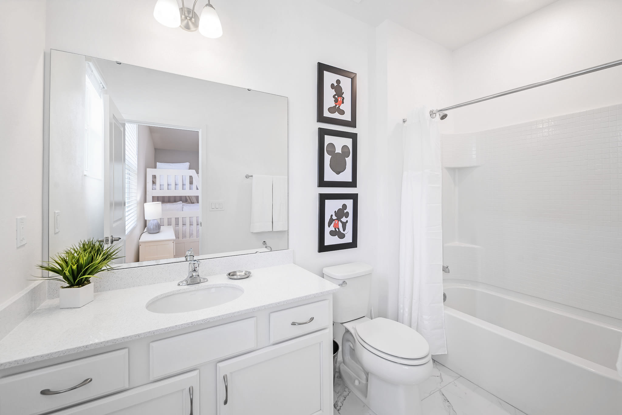 Clean white bathroom with a single vanity, large mirror reflecting a bunk bed room, a toilet, and a bathtub with a shower. Mickey Mouse framed artwork adorns the wall.
