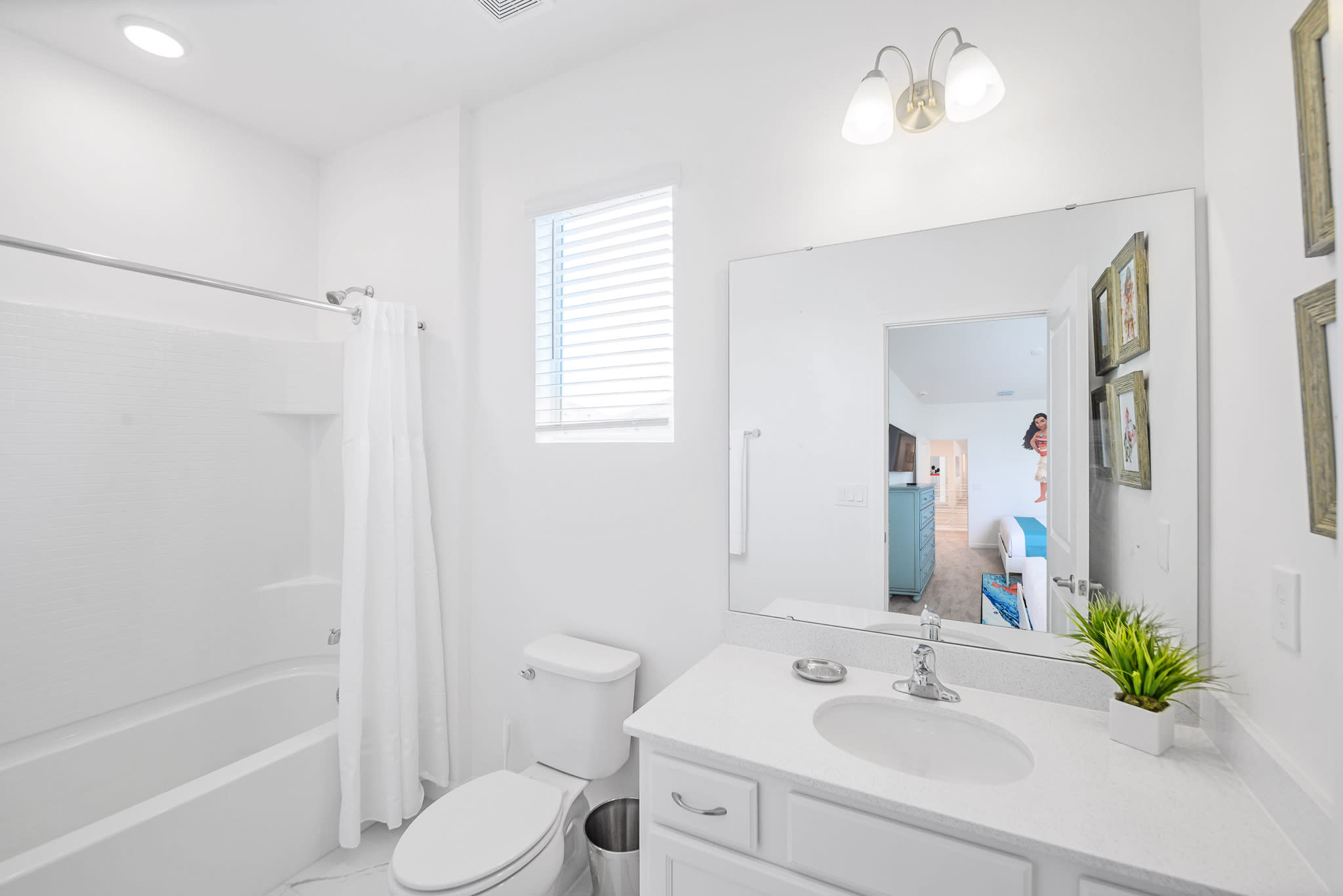 Bright white bathroom featuring a single vanity with a large mirror, a toilet, and a bathtub with a shower and white curtain. A window with blinds is visible above the toilet.