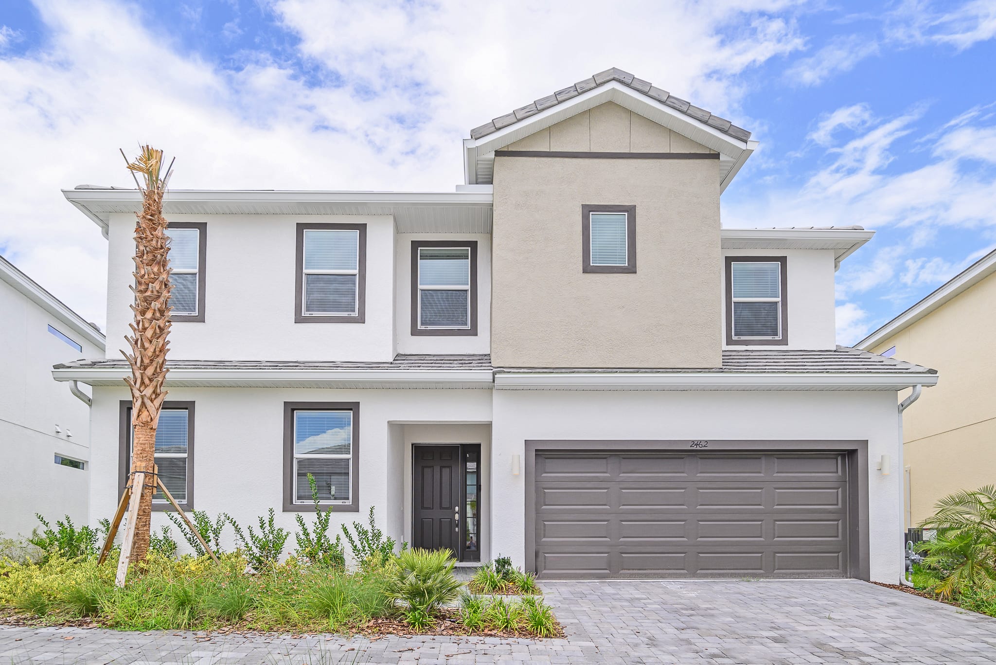 Modern two-story house with a white and beige facade, dark gray garage door, and a dark brown front door. Landscaping includes small shrubs and a tall palm tree.