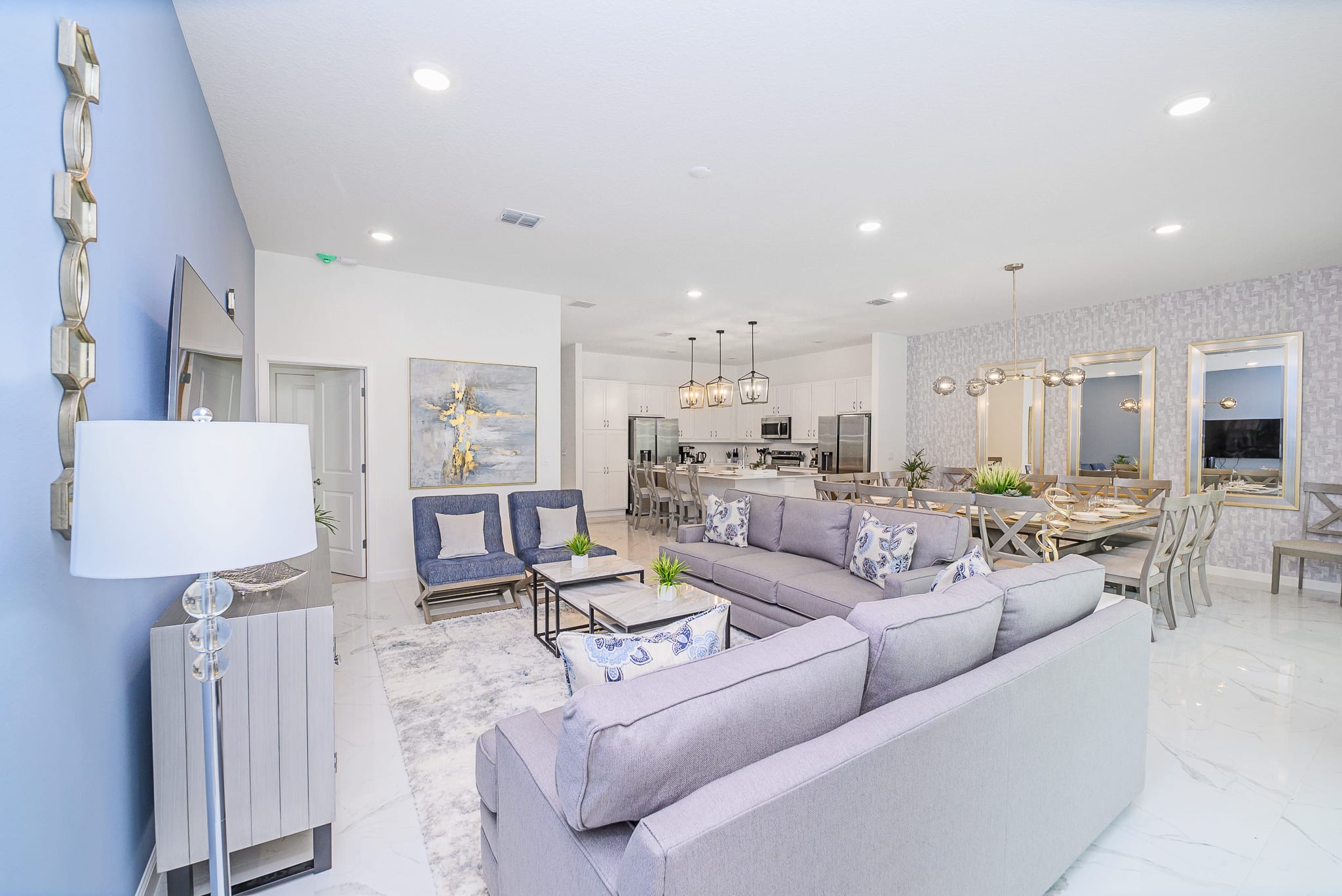 An expansive open-concept living area with light grey sofas and armchairs, a marble coffee table, and a large area rug, flowing into a modern kitchen and dining space with white cabinetry and light flooring.
