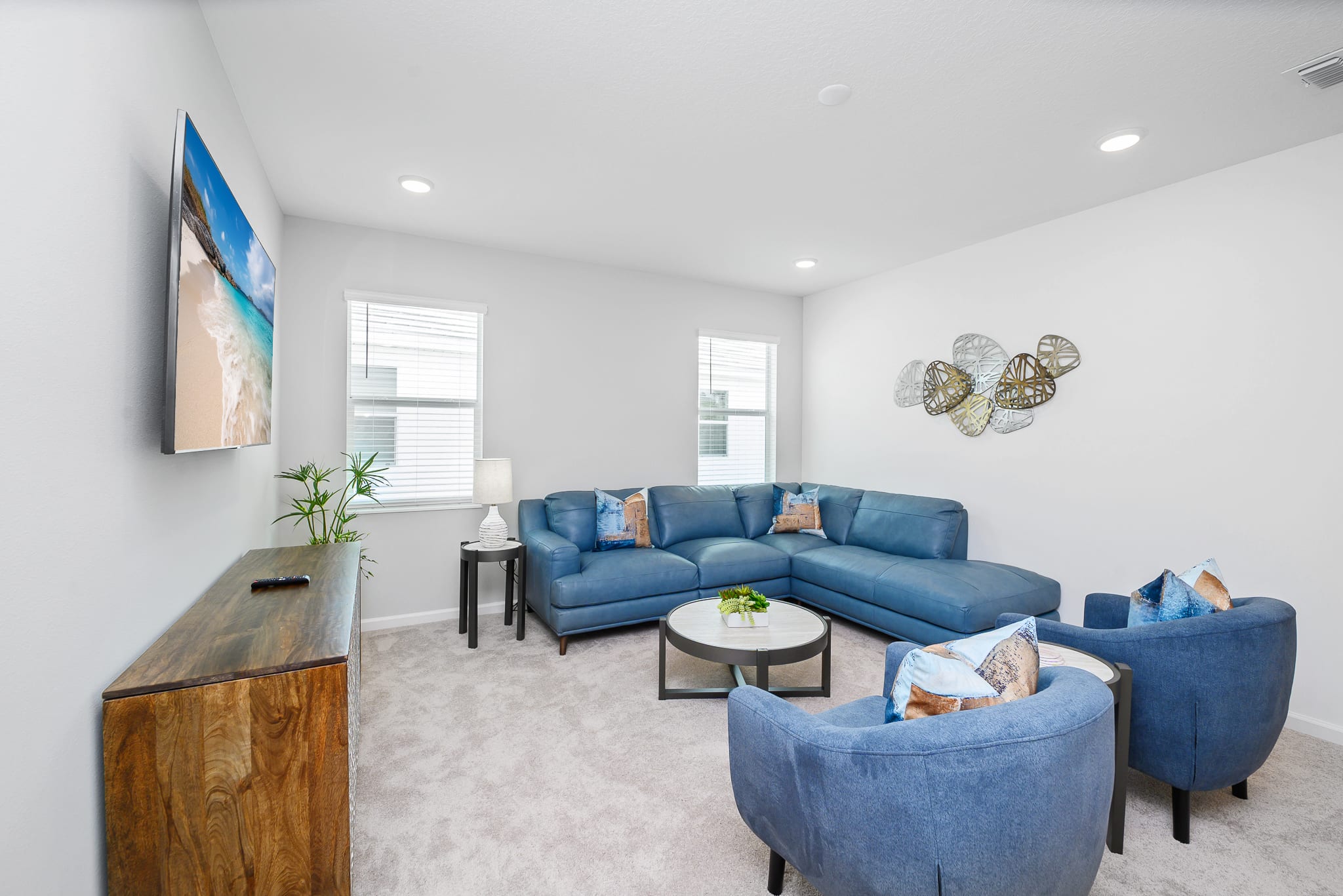 Cozy living room with a large blue leather sectional sofa and two matching armchairs arranged around a round coffee table. A wall-mounted TV displays a beach scene, and decorative metal wall art adorns the white wall above the sofa.