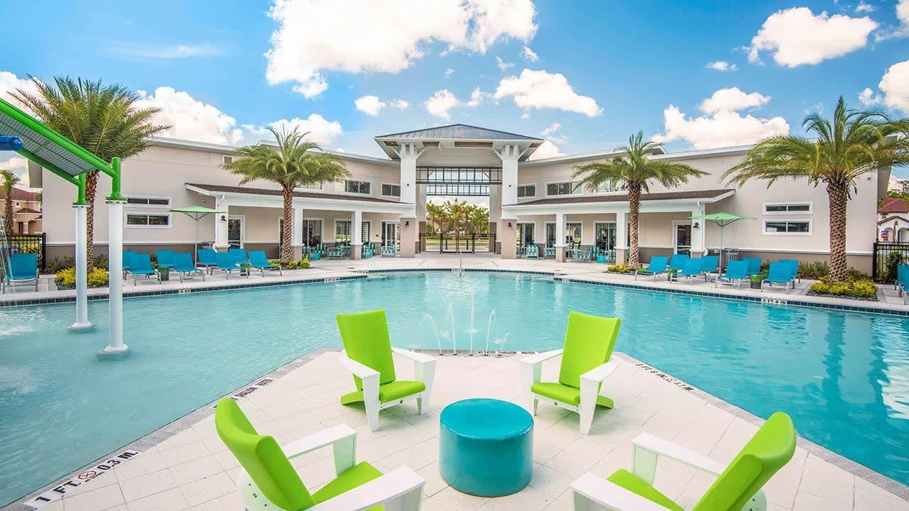Expansive communal swimming pool area with a large pool, numerous lounge chairs, and a modern clubhouse building in the background under a bright blue sky.
