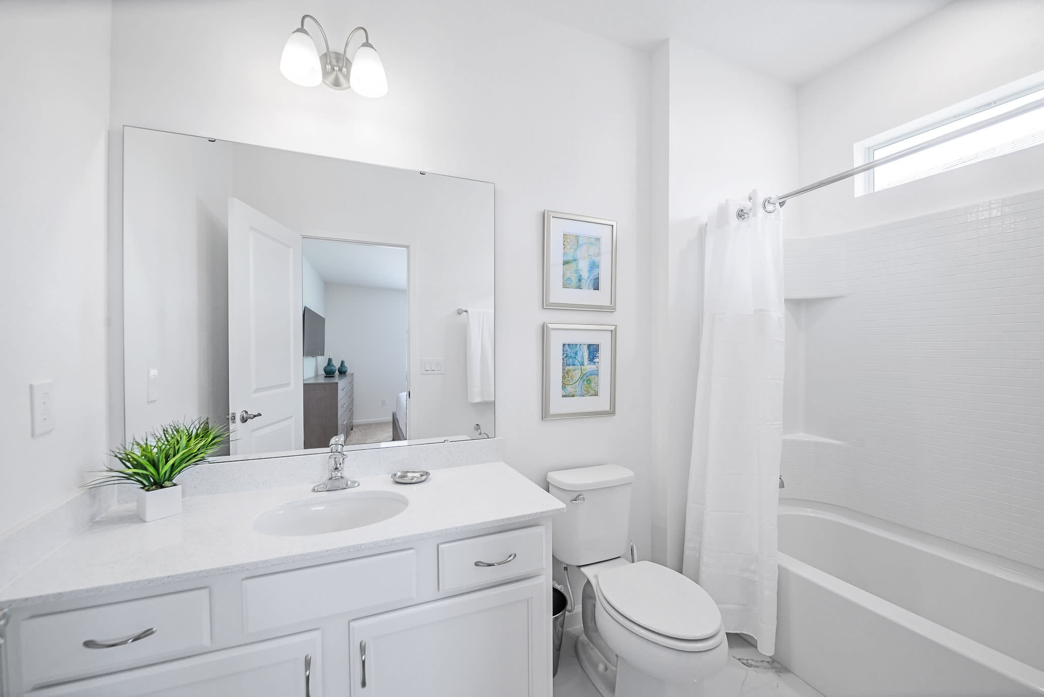 Bright white bathroom with a single vanity, large mirror, toilet, and a bathtub with a shower. The shower features white subway tile, and the room is accented with framed artwork and a small potted plant.
