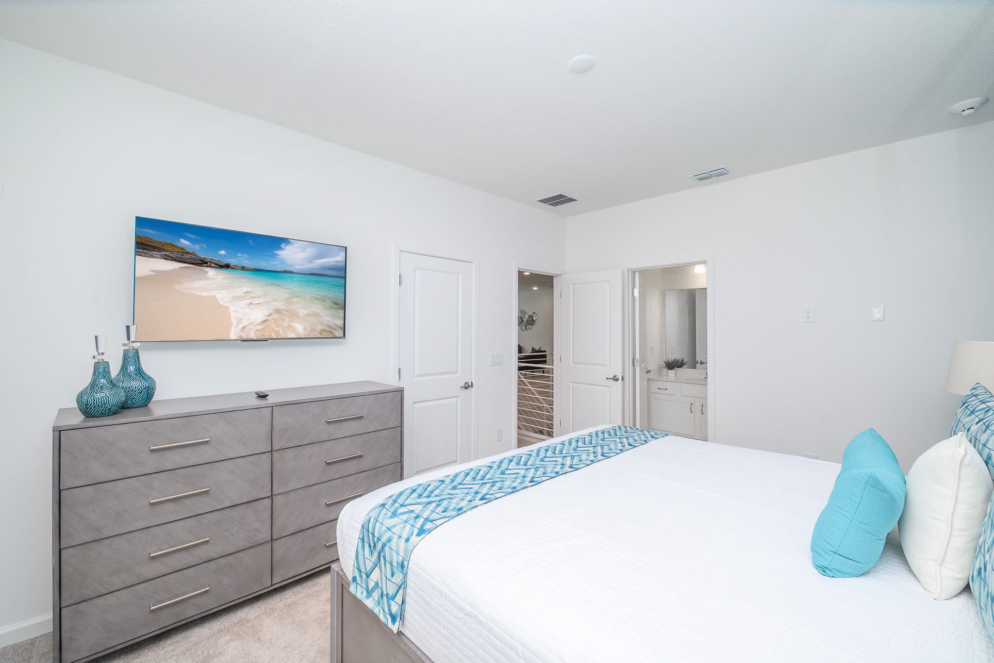 Another view of bedroom-2, showcasing a large grey dresser with a wall-mounted TV and teal decorative vases. White doors lead to other areas, maintaining the room's clean and modern aesthetic.