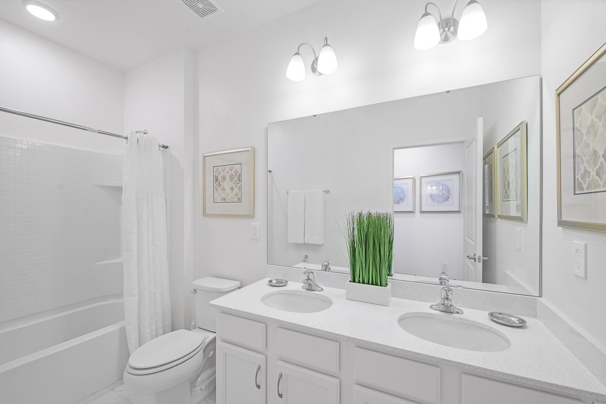 Spacious white bathroom featuring a double vanity with two sinks, white countertops, and chrome faucets. A large mirror spans the width of the vanity, reflecting framed artwork and a bathtub/shower combo.