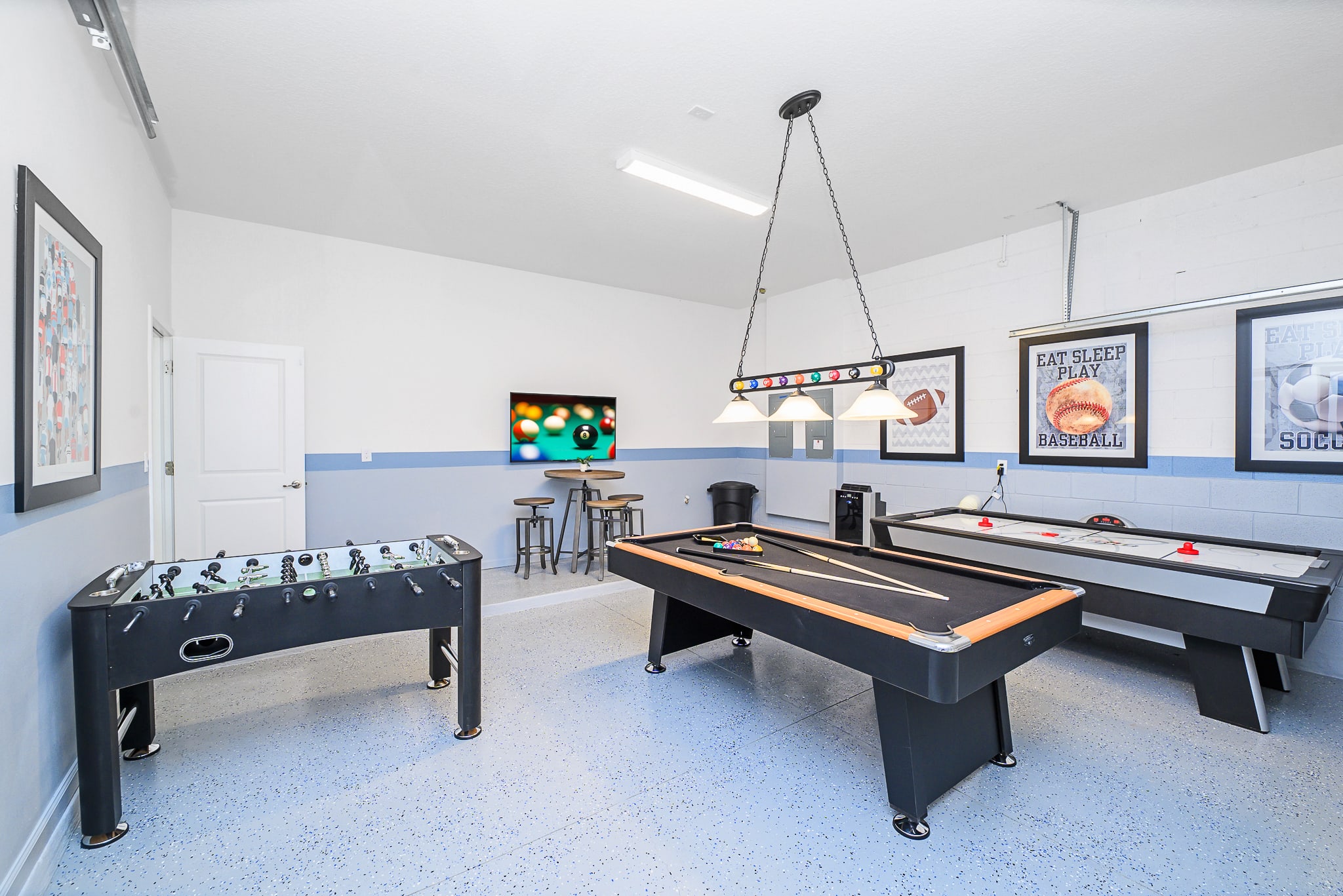 A spacious game room, likely a converted garage, features a foosball table, a pool table with an overhead light, and an air hockey table. A high-top table with stools and a wall-mounted TV showing a game of pool complete the entertainment space, set against a two-tone blue and white wall.