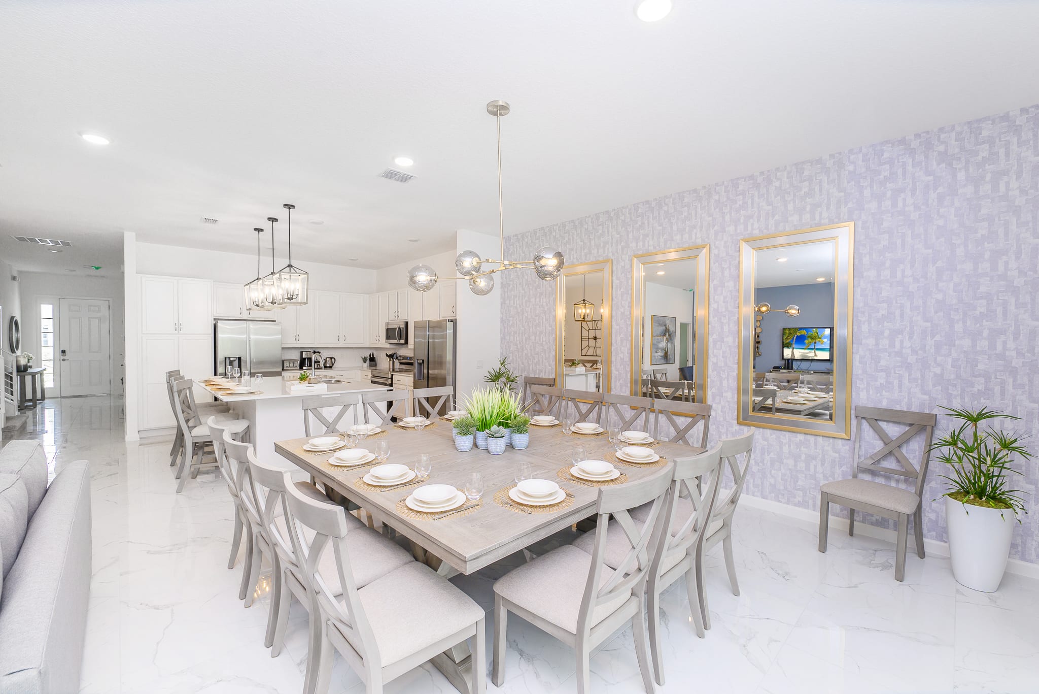 A bright dining room with a large square light wood table and grey upholstered chairs, illuminated by a contemporary chandelier, with decorative mirrors on the wall and the kitchen visible in the background.