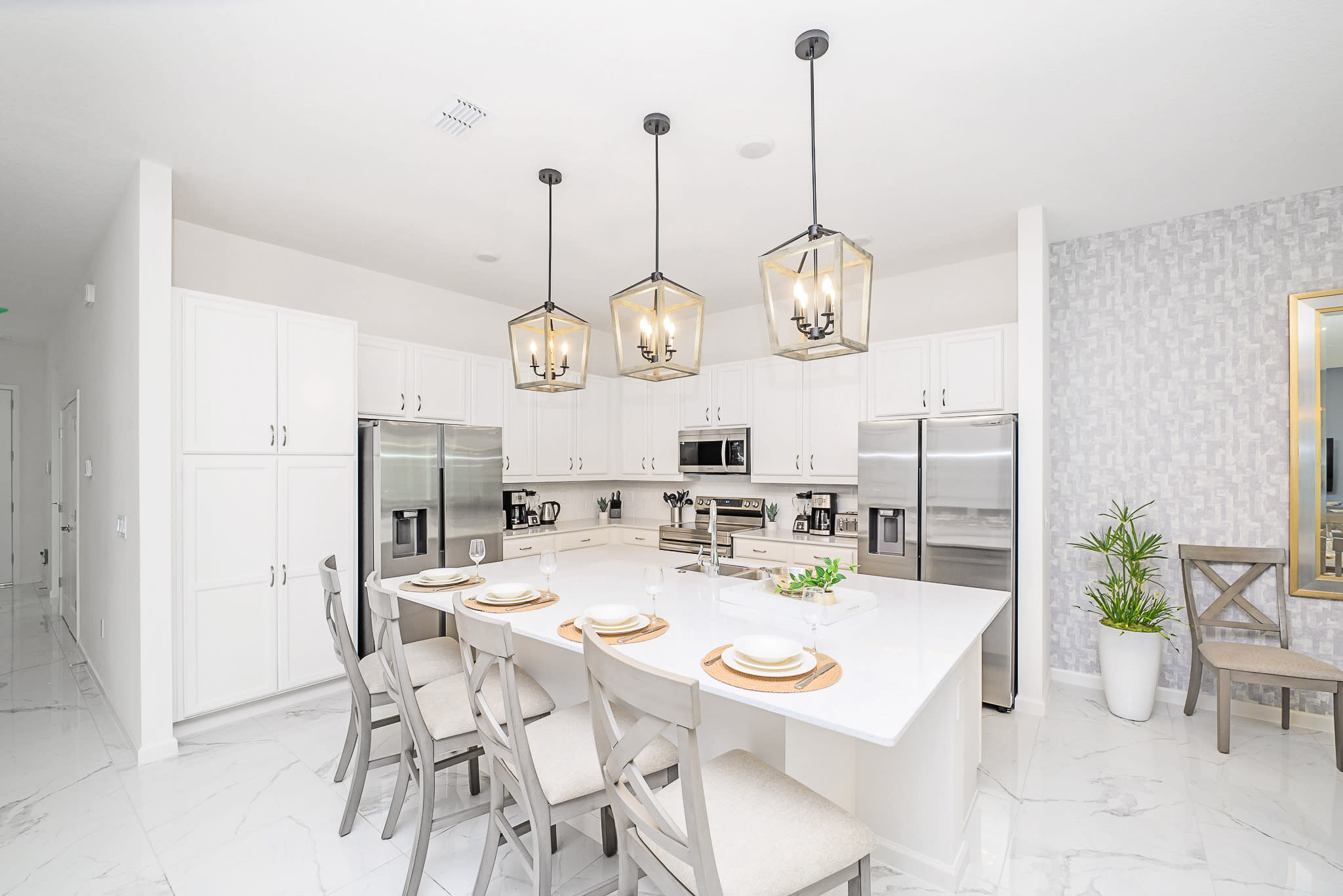 A modern white kitchen featuring a large island with seating for four, stainless steel appliances including two refrigerators, white shaker cabinets, and three contemporary pendant lights above the island.