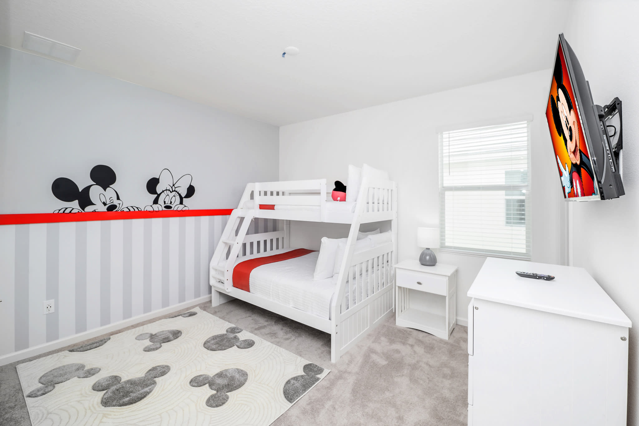 Children's bedroom with a white wooden bunk bed, Mickey Mouse themed wall decor, and a matching rug. A wall-mounted TV and a small nightstand with a lamp are also present.