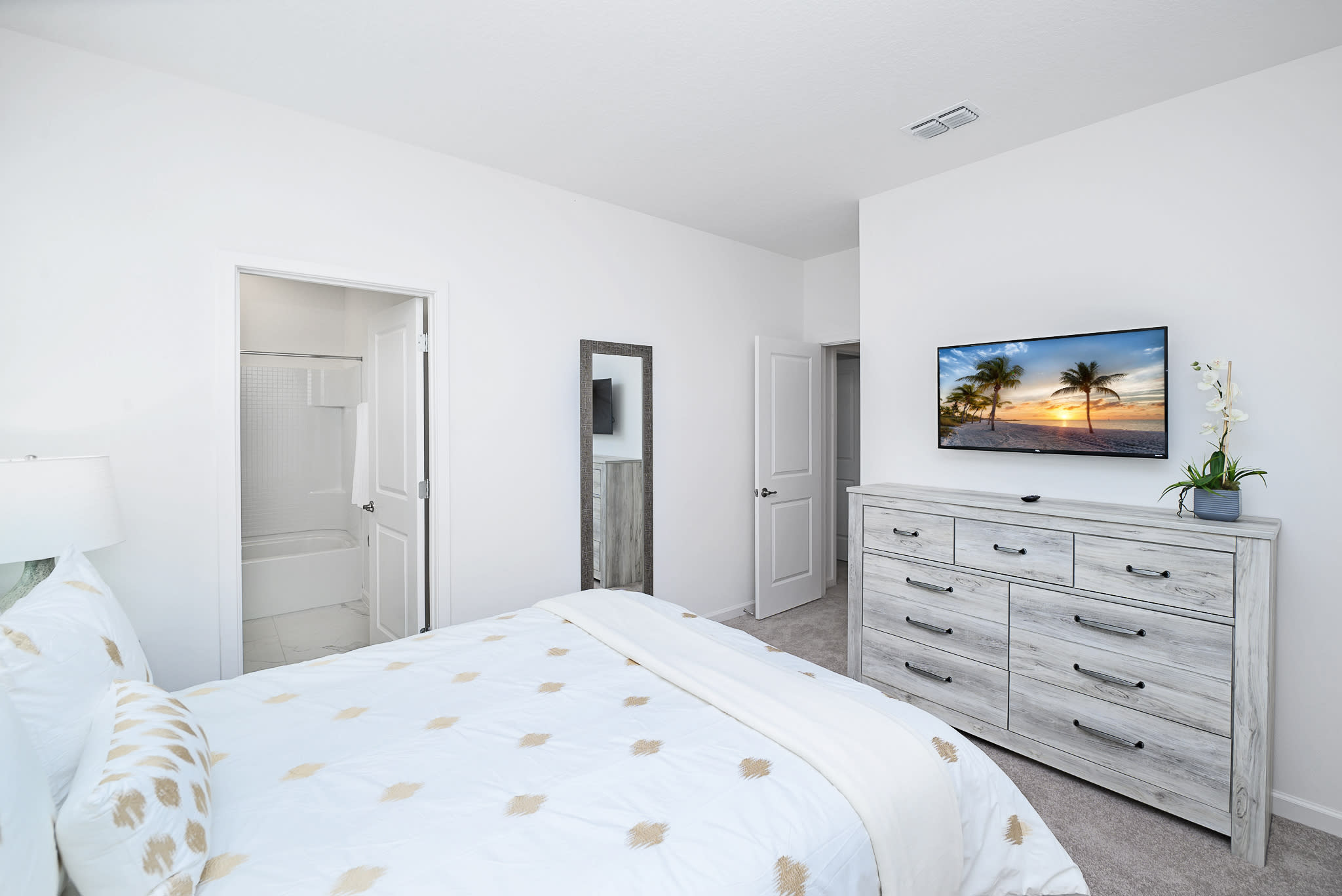 View of a bedroom featuring a wall-mounted TV above a light wood dresser with multiple drawers. A full-length mirror stands near the doorway leading to a bathroom with a tub/shower combo.