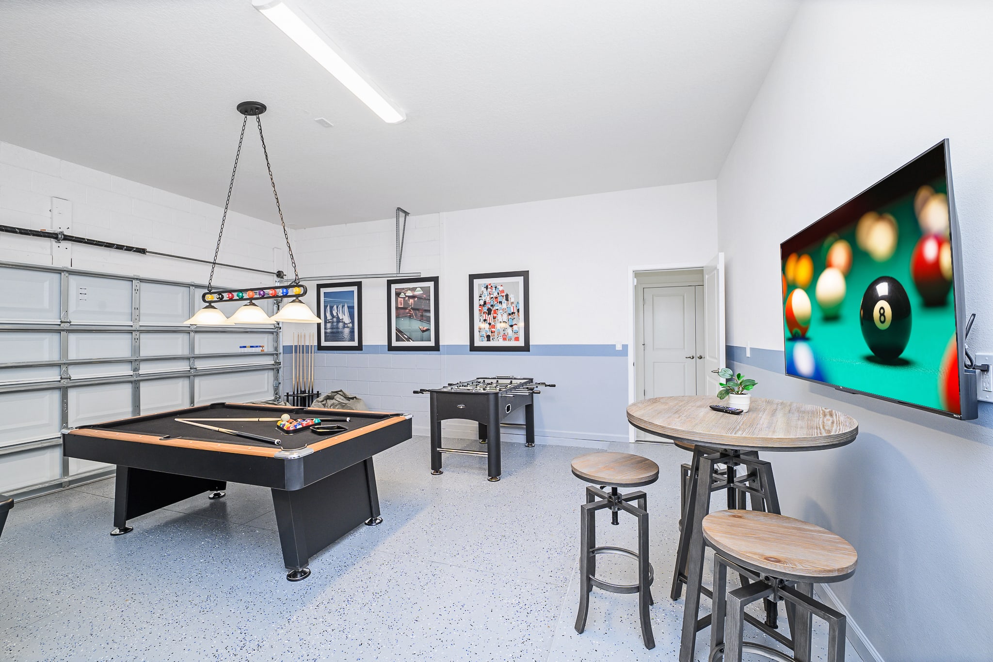 The game room, set in a converted garage, offers a variety of entertainment with a pool table, foosball table, and a high-top table with stools. A large wall-mounted TV displays a game, and decorative framed art adorns the two-tone walls.
