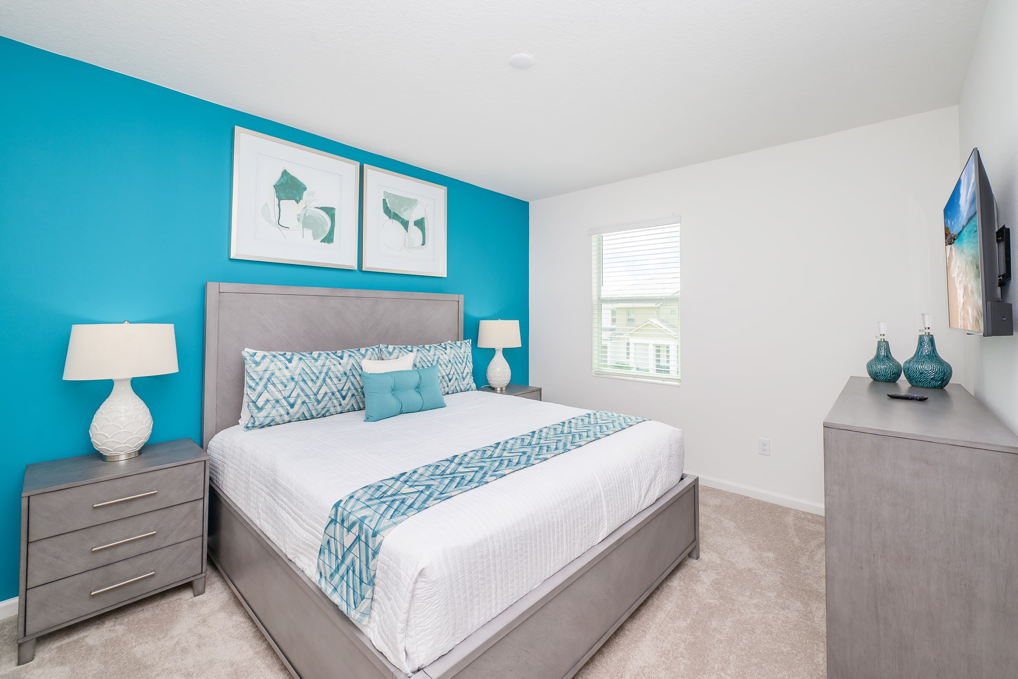 Modern bedroom with a vibrant teal accent wall behind a grey upholstered bed, flanked by matching grey nightstands with lamps. The bedding features a blue and white geometric pattern, enhancing the contemporary feel.