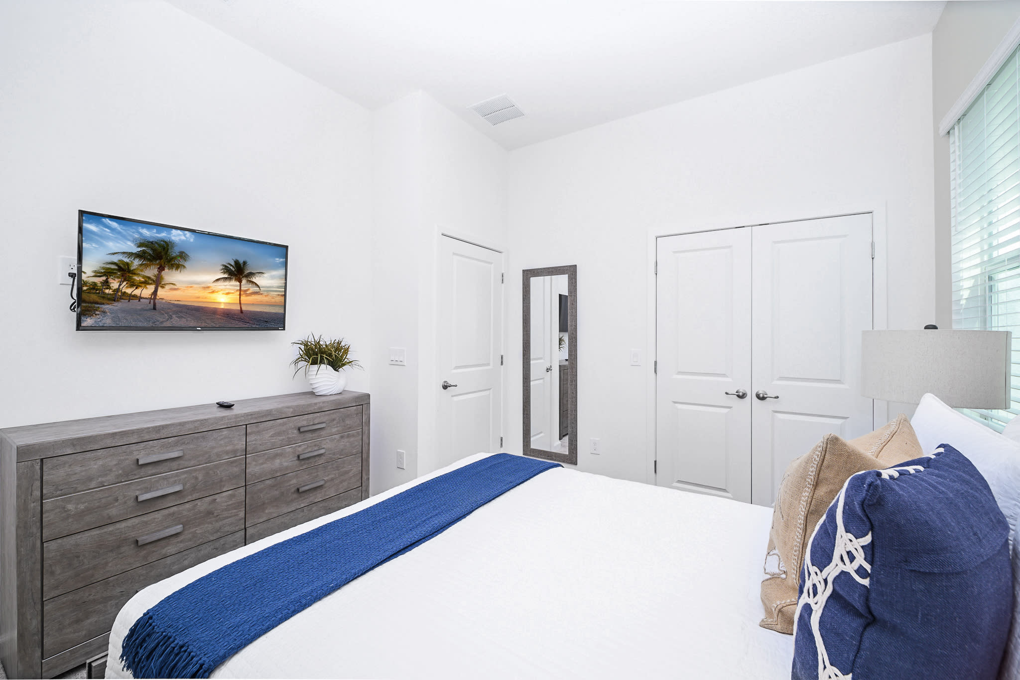 View of bedroom-4 showing a grey wooden dresser with a wall-mounted TV and a potted plant. White closet doors and a full-length mirror are visible, maintaining a light and airy feel in the room.