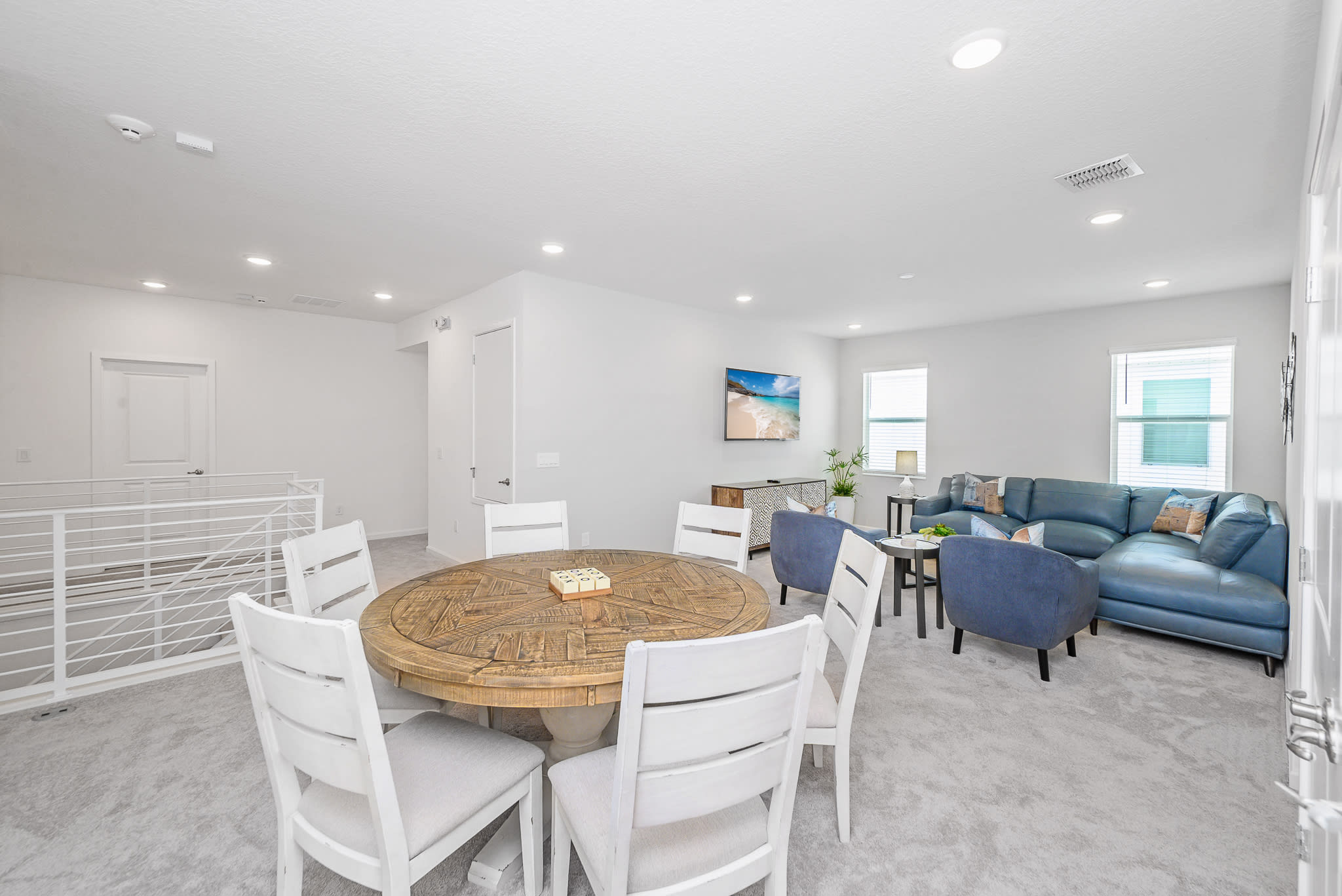 Expansive upstairs common area featuring a blue sectional sofa and armchairs in the living space, and a round wooden dining table with white chairs. A white railing indicates an open view to a lower level.