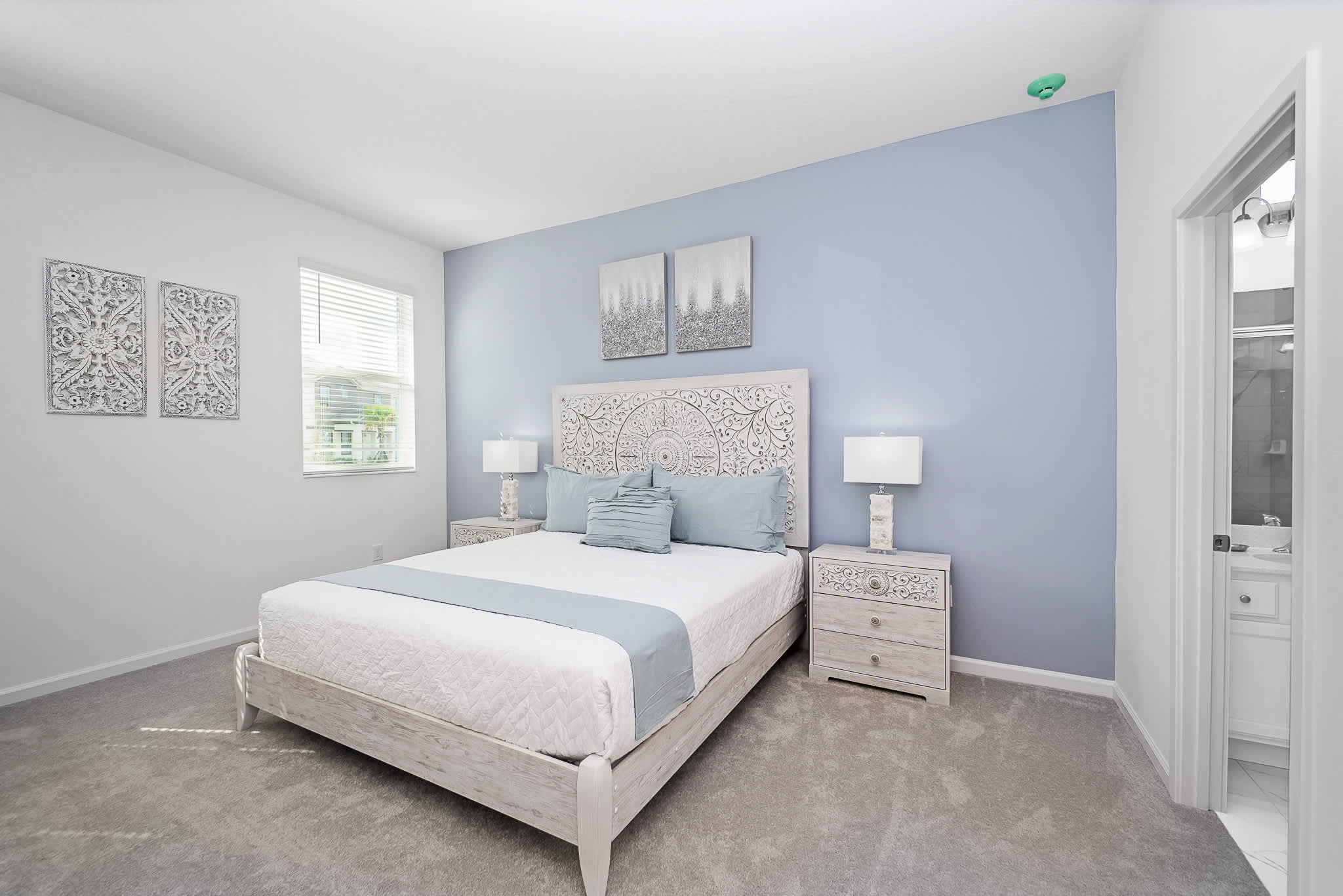 Serene bedroom featuring a light blue accent wall behind a white bed with an intricately carved headboard. Two matching nightstands with lamps flank the bed, and a window with blinds provides natural light.