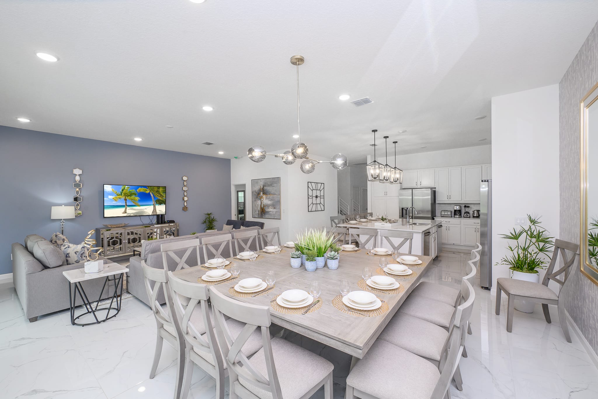 A spacious dining area featuring a large square wooden table with seating for many, set with place settings, under a modern chandelier, with the living room and kitchen visible in the background.