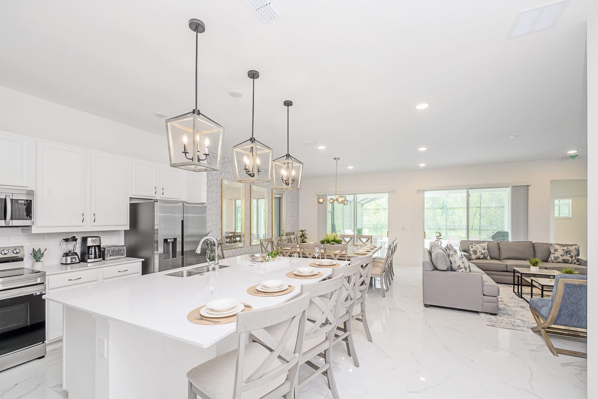 A spacious, open-concept kitchen with a large white island and bar seating, white cabinetry, and stainless steel appliances, seamlessly connecting to the dining and living areas, all with light tile flooring.
