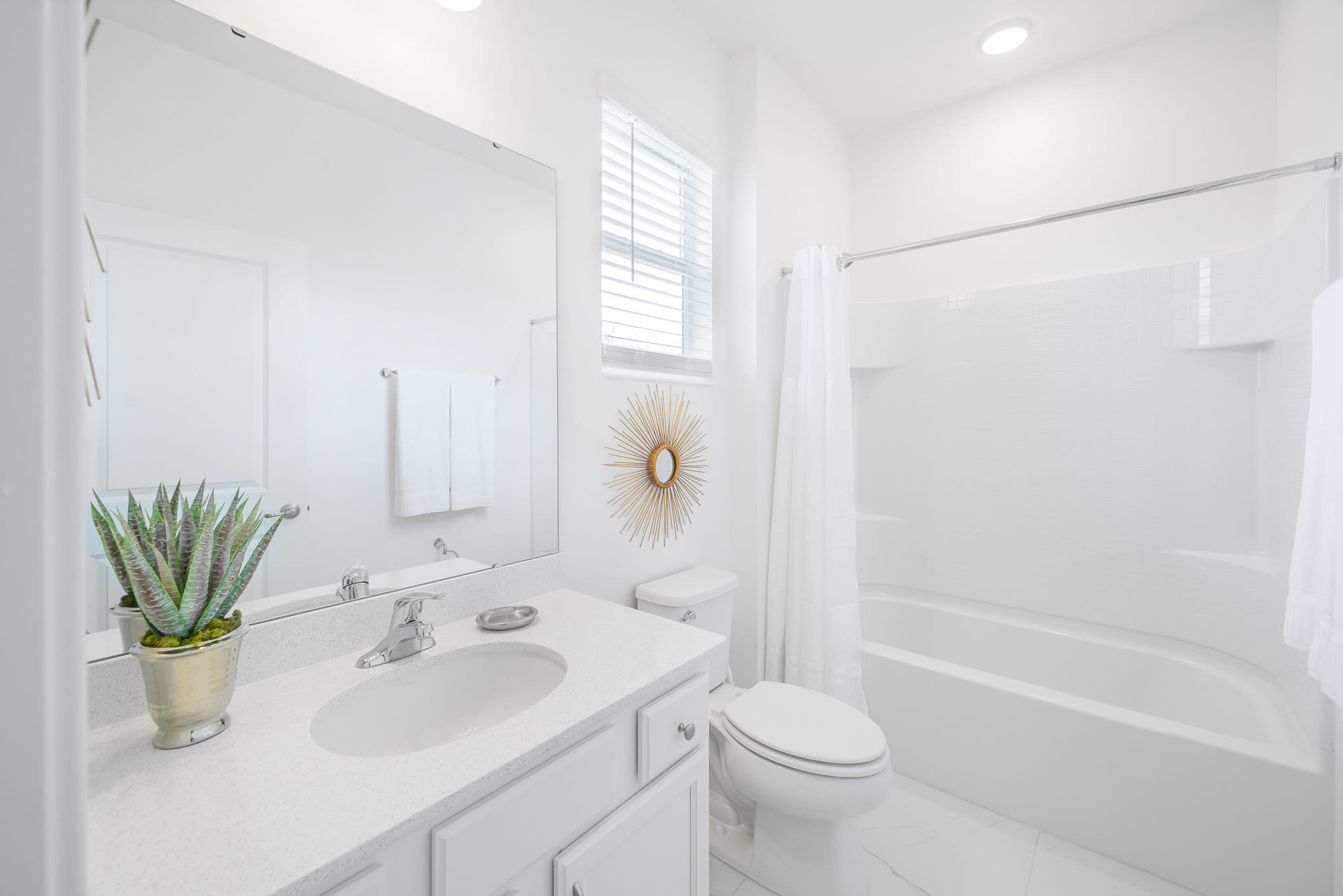 Modern white bathroom with a single vanity, white countertop, and a large mirror. A window with blinds is positioned above the toilet, and a gold sunburst mirror decorates the wall.