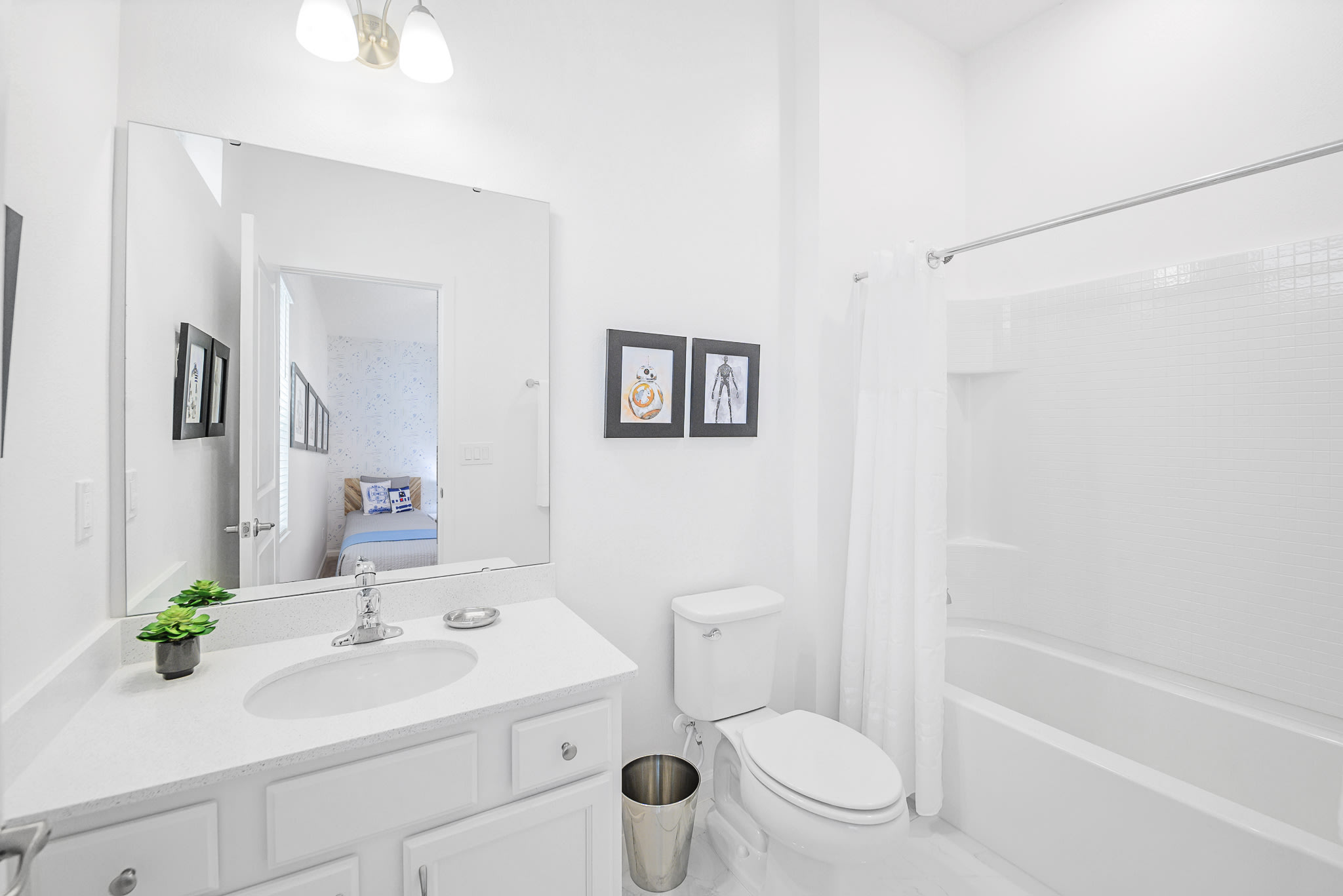 Clean white bathroom with a single vanity, white countertop, and a large mirror reflecting the Star Wars-themed bedroom. Two framed artworks hang above the toilet next to a bathtub/shower combo.