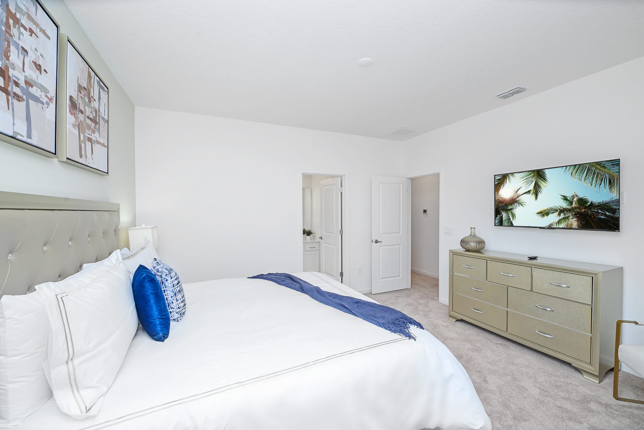 View of bedroom-3 showing a gold-toned dresser with a wall-mounted TV and a decorative vase. Two white doors lead to other areas, and the room maintains a light, neutral color palette with plush carpeting.