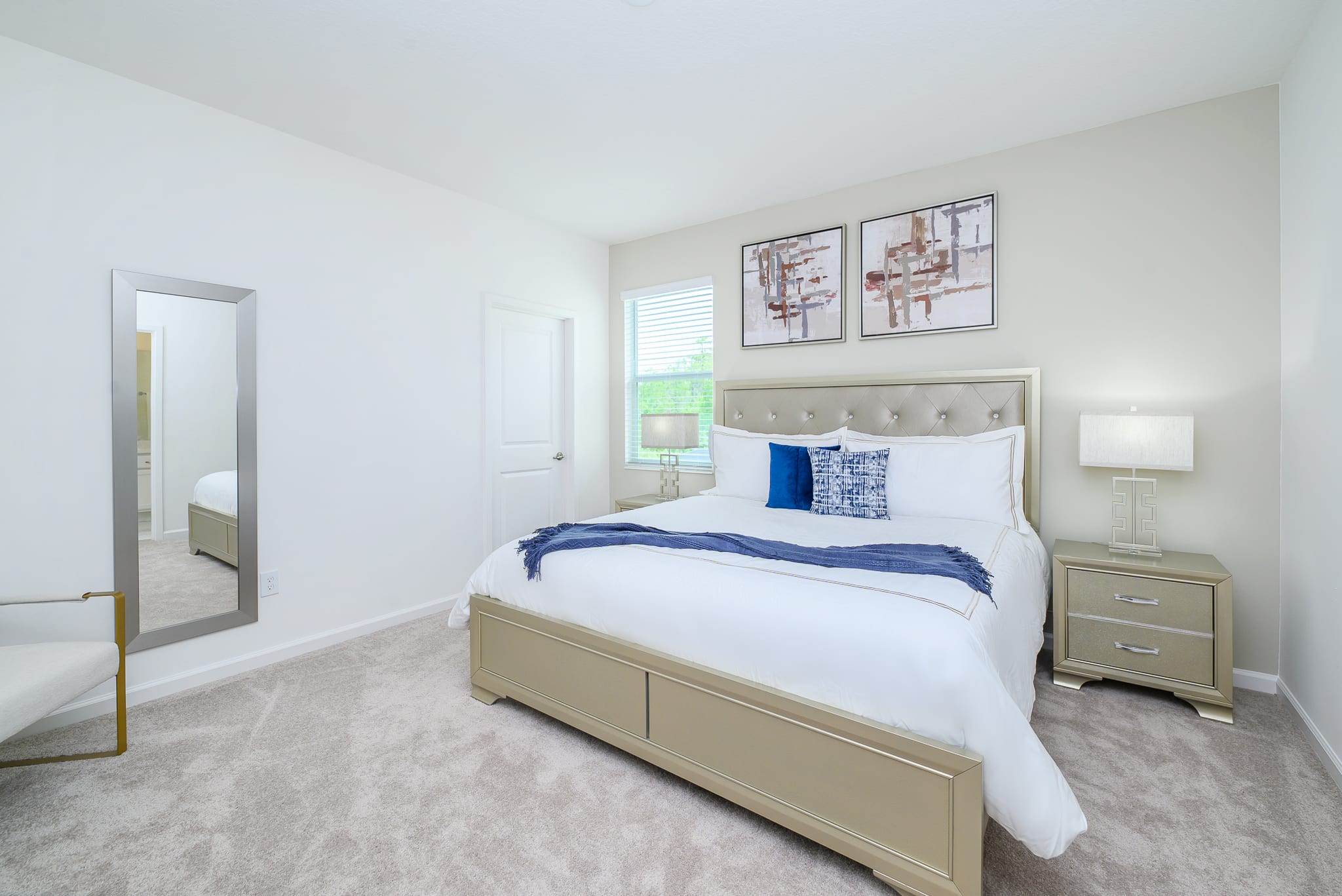 Elegant bedroom featuring a tufted upholstered headboard on a champagne-colored bed frame, flanked by matching nightstands with geometric lamps. A full-length mirror and a small accent chair add to the sophisticated decor.