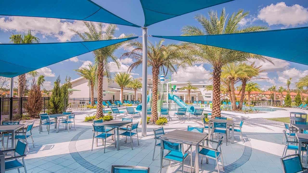 An expansive outdoor dining area is shaded by large blue fabric sails, featuring numerous square tables with matching blue chairs. Palm trees are interspersed throughout the paved area, with a communal swimming pool and water play features visible in the background.