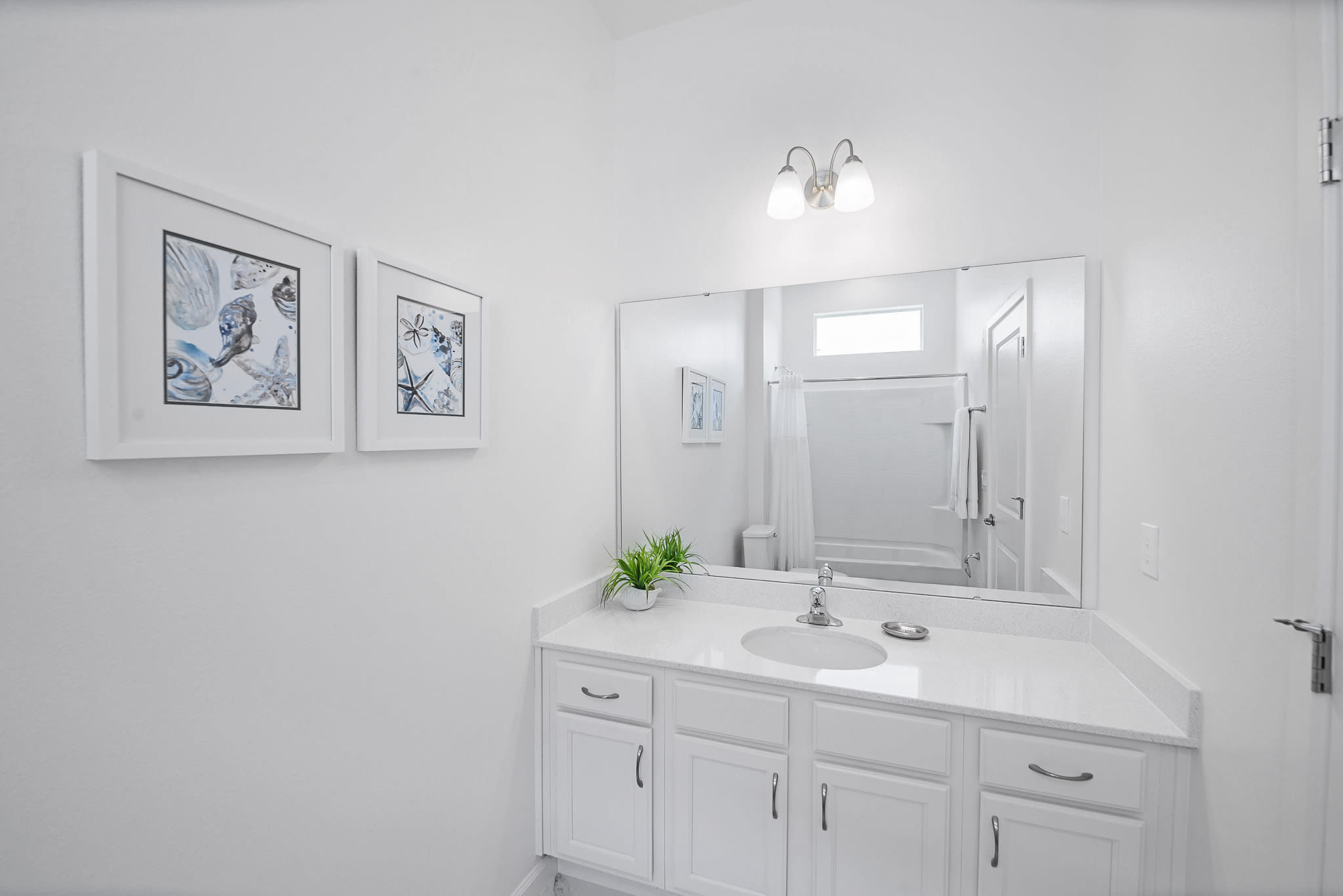 Bright white bathroom featuring a single vanity with a white countertop, chrome faucet, and a large mirror reflecting a bathtub/shower combo. Two framed coastal-themed artworks adorn the wall.