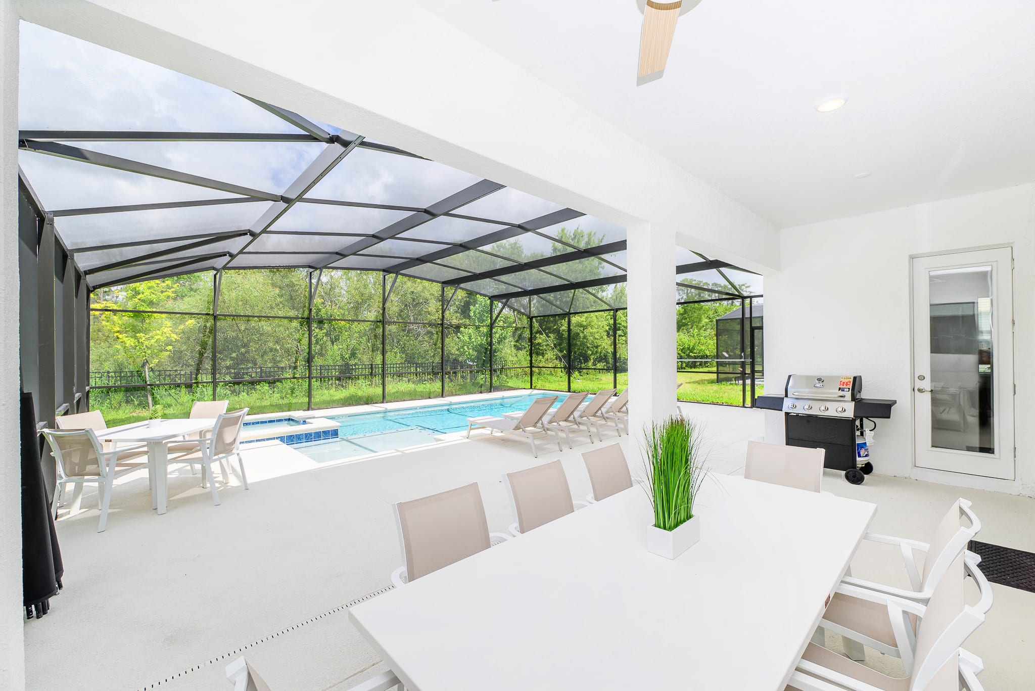 This view from the covered patio highlights a large outdoor dining table and a BBQ grill, with the screened-in private pool and hot tub visible just beyond. The bright white patio extends to the pool deck, offering ample space for relaxation and outdoor entertaining.
