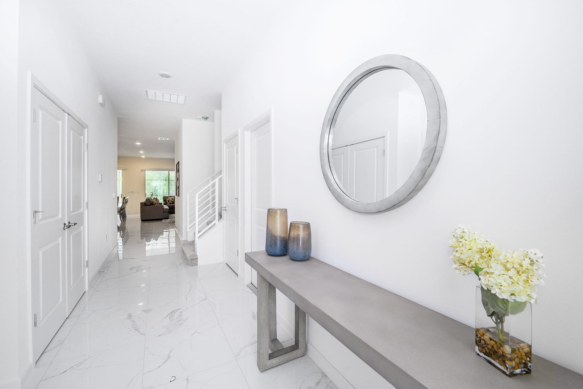 Bright and modern foyer featuring white marble-look tile flooring, a grey console table with decorative vases, and a large round mirror on a white wall. A staircase with white railings is visible in the background.