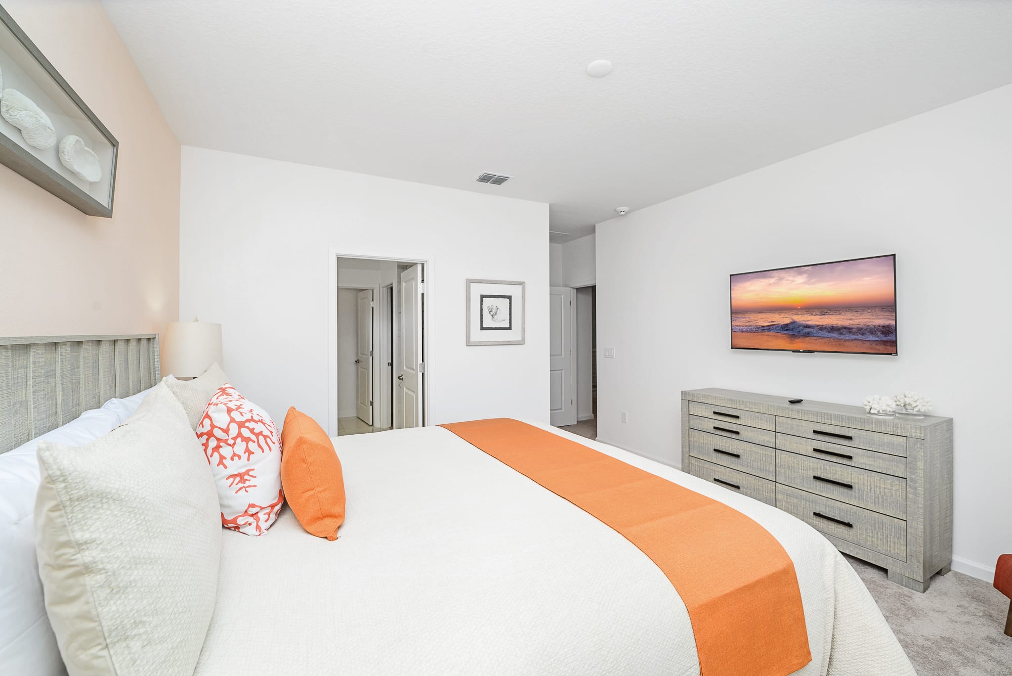 Comfortable bedroom with a grey wooden bed frame and orange accent pillows and bed runner. A wall-mounted TV is positioned above a matching grey dresser, and an open doorway leads to an en-suite bathroom.