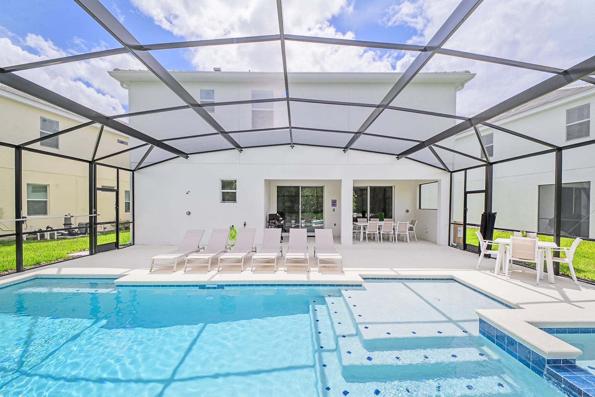 The expansive screened-in outdoor pool area showcases a sparkling blue swimming pool with integrated steps and a hot tub, surrounded by a clean white patio. Several lounge chairs are arranged for sunbathing, with a covered patio area featuring an outdoor dining set and a grill visible in the background.