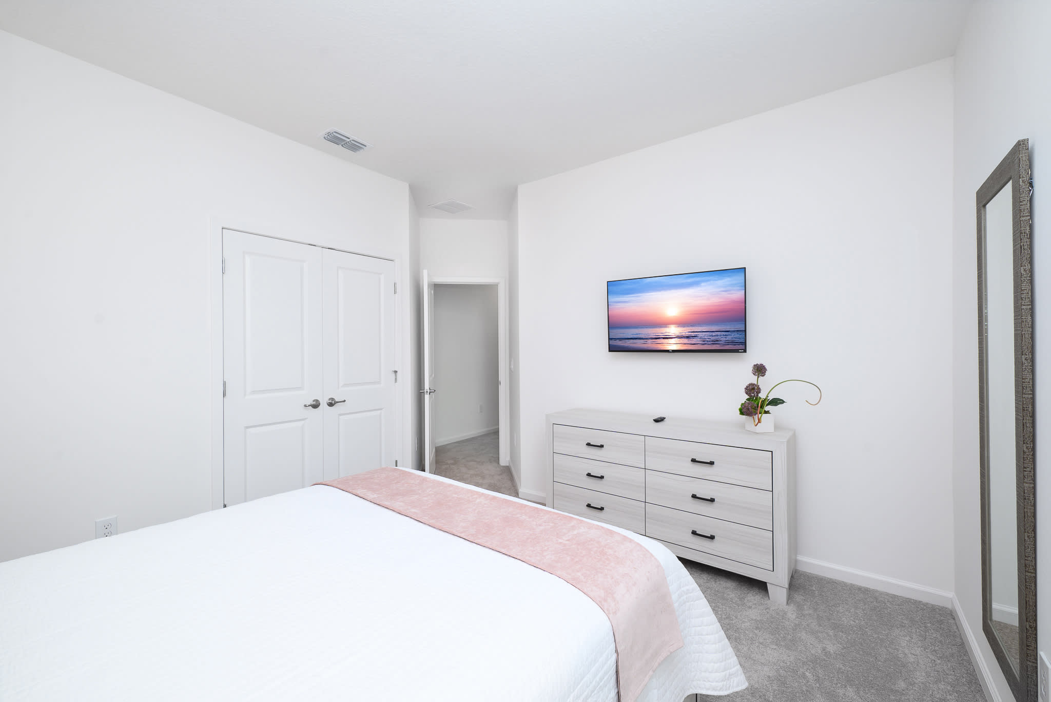 Another view of the bedroom, showcasing a wall-mounted TV above a white dresser with black handles. A full-length mirror leans against the wall, and a closet with bifold doors is visible.