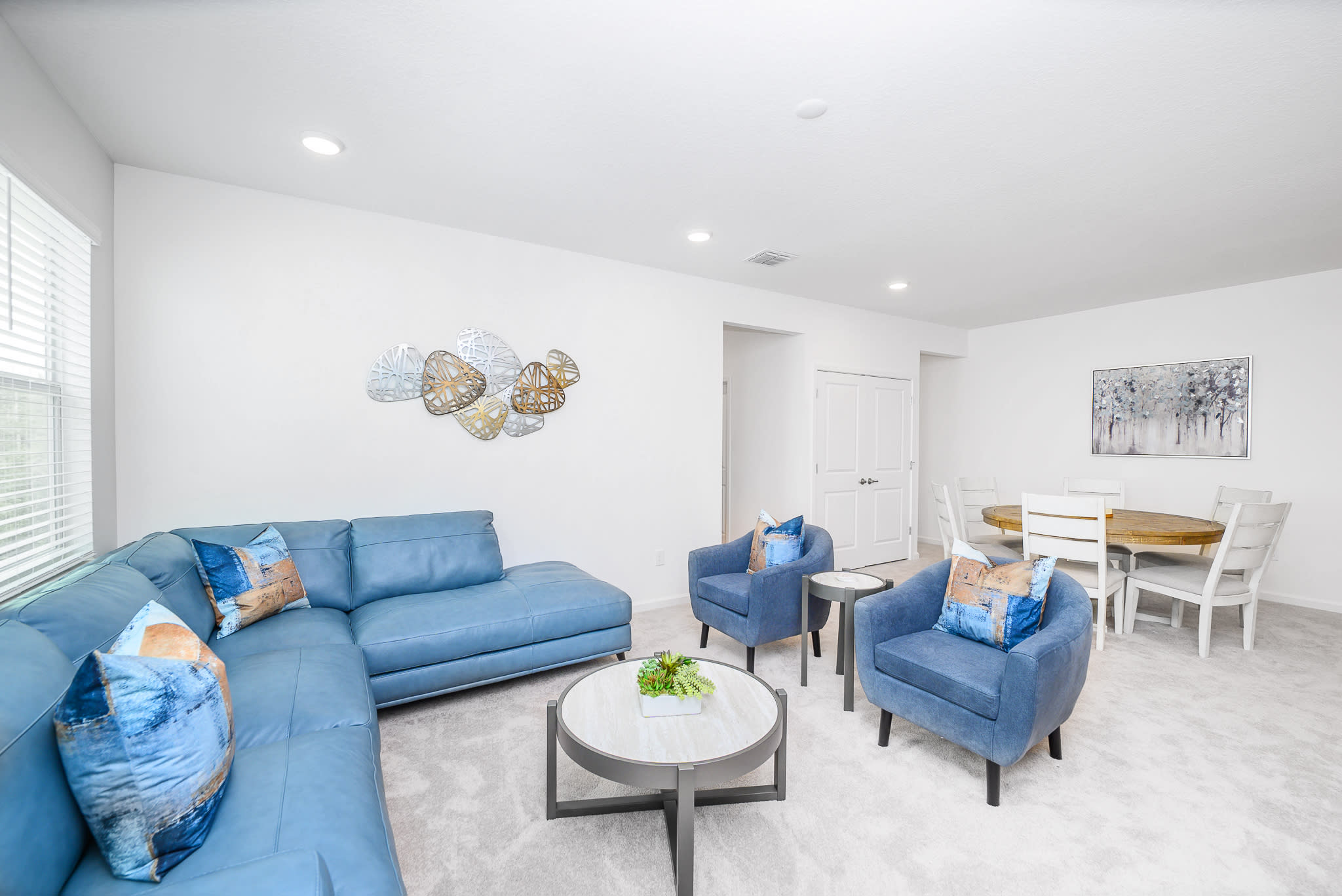 Spacious open-concept living area featuring a blue sectional sofa and armchairs, a round coffee table, and decorative metal wall art. A dining area with a round wooden table and white chairs is visible in the background.