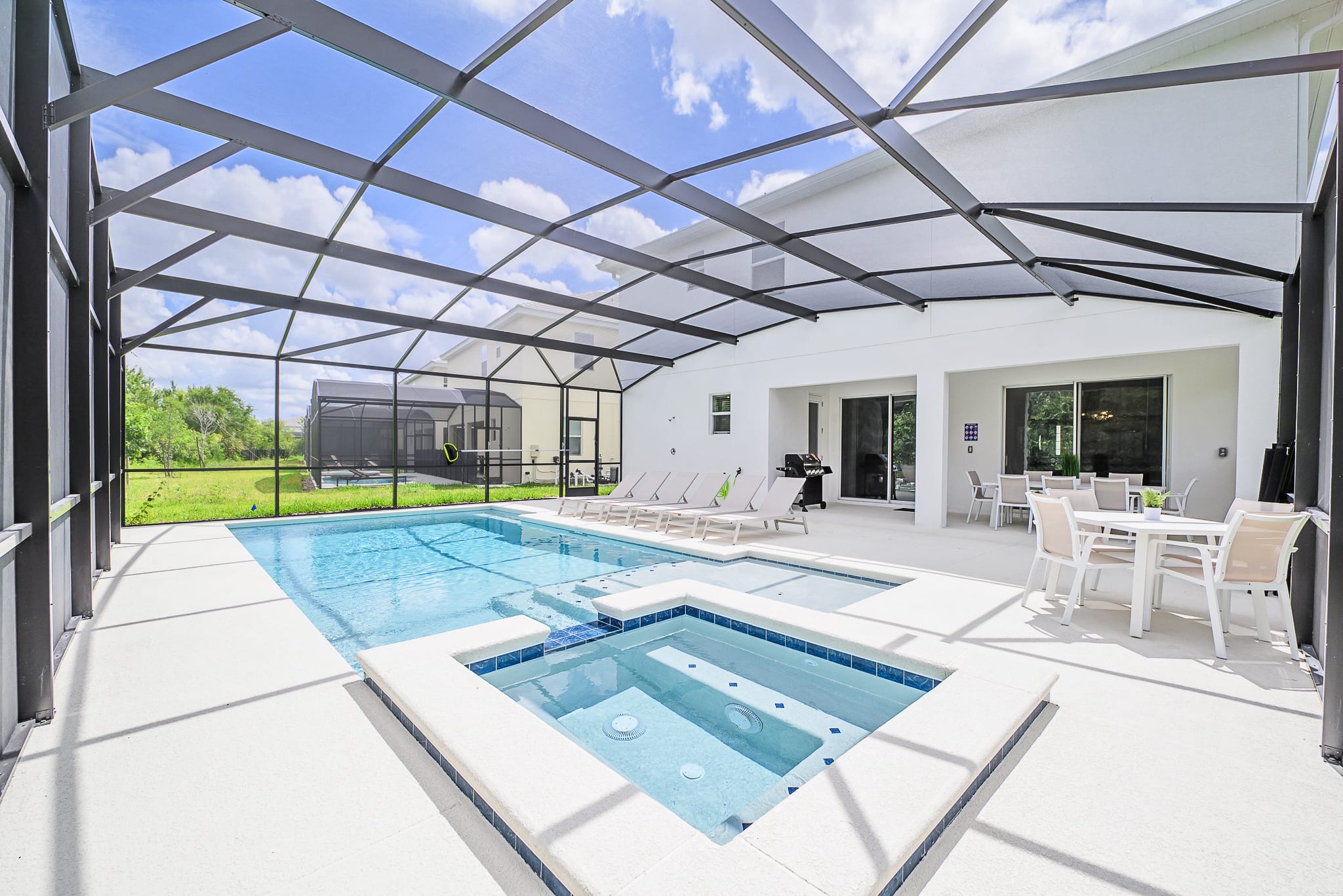 A large screened-in outdoor pool area features a rectangular swimming pool with an integrated hot tub, surrounded by a white patio with lounge chairs and an outdoor dining table. The enclosure provides shade and protection, with a view of a grassy backyard and another screened enclosure in the distance.