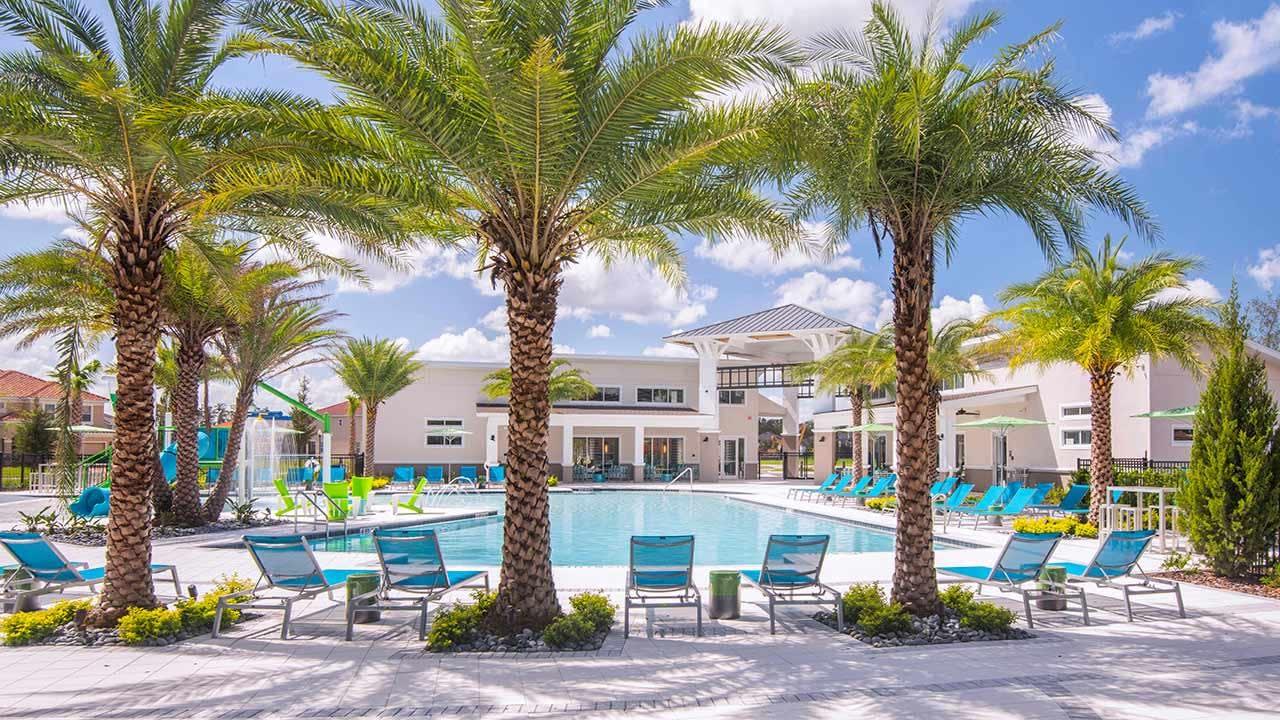 A large, resort-style outdoor swimming pool is surrounded by numerous teal lounge chairs and tall palm trees under a bright blue sky. A modern clubhouse building with a white exterior and large windows stands in the background, suggesting a communal amenity space.