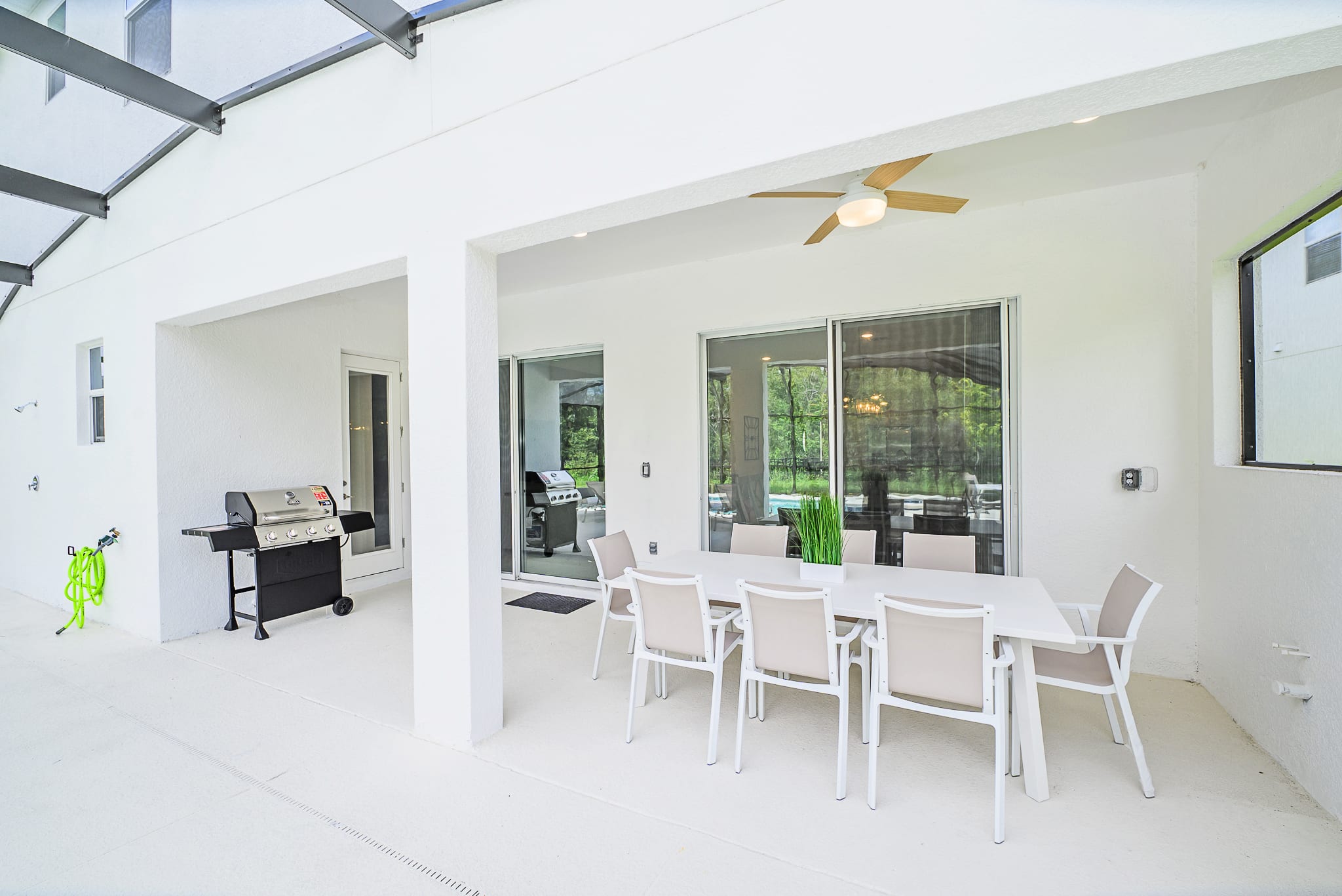 A spacious covered patio area features a large white outdoor dining table with seating for eight, positioned next to a BBQ grill. Sliding glass doors lead into the house, while a ceiling fan provides comfort, and the screened pool area is visible beyond.