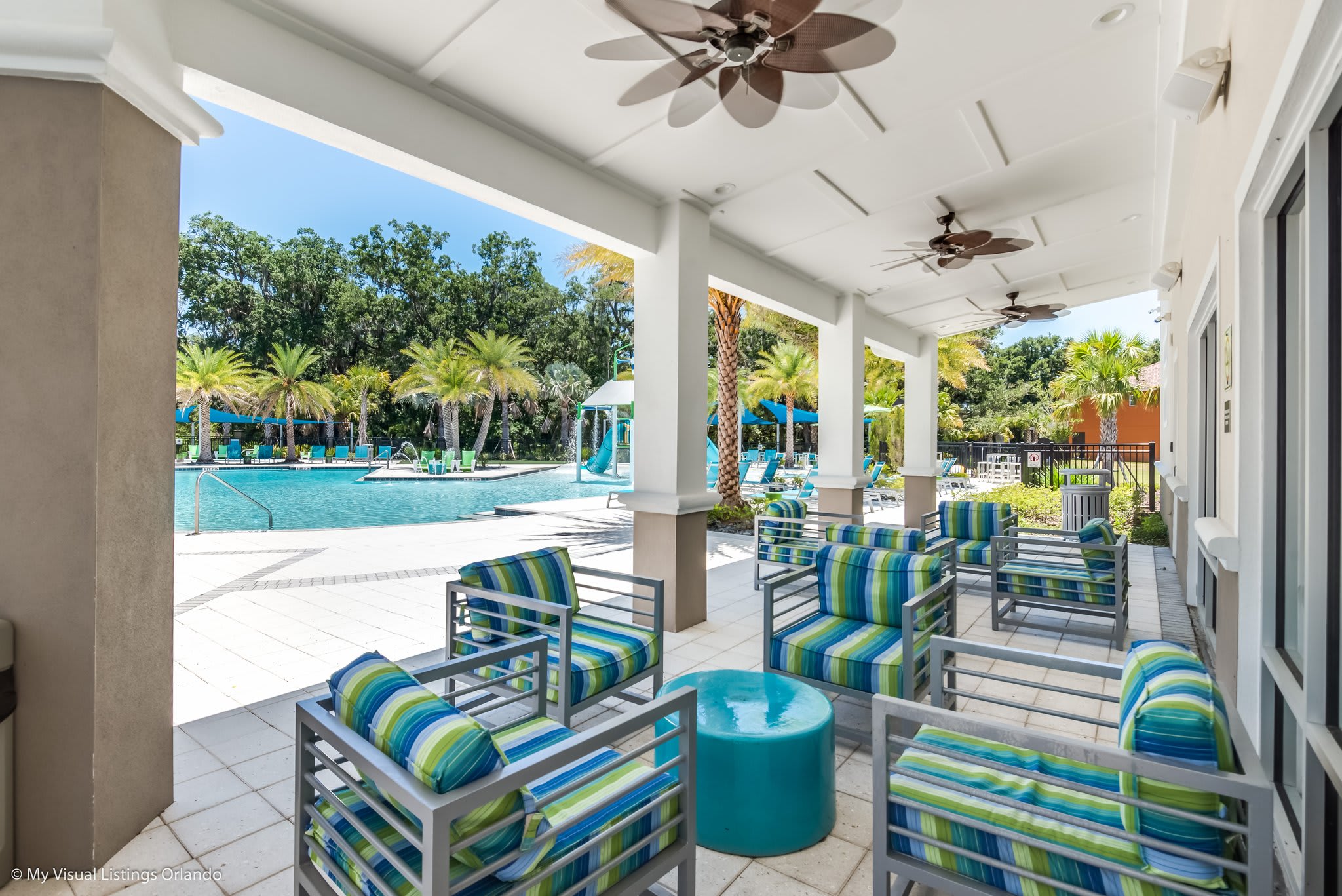 A covered outdoor patio features comfortable modern seating with striped blue and green cushions, arranged around a round teal table. Beyond the patio, a large swimming pool is visible, surrounded by palm trees and lounge chairs under a clear blue sky.