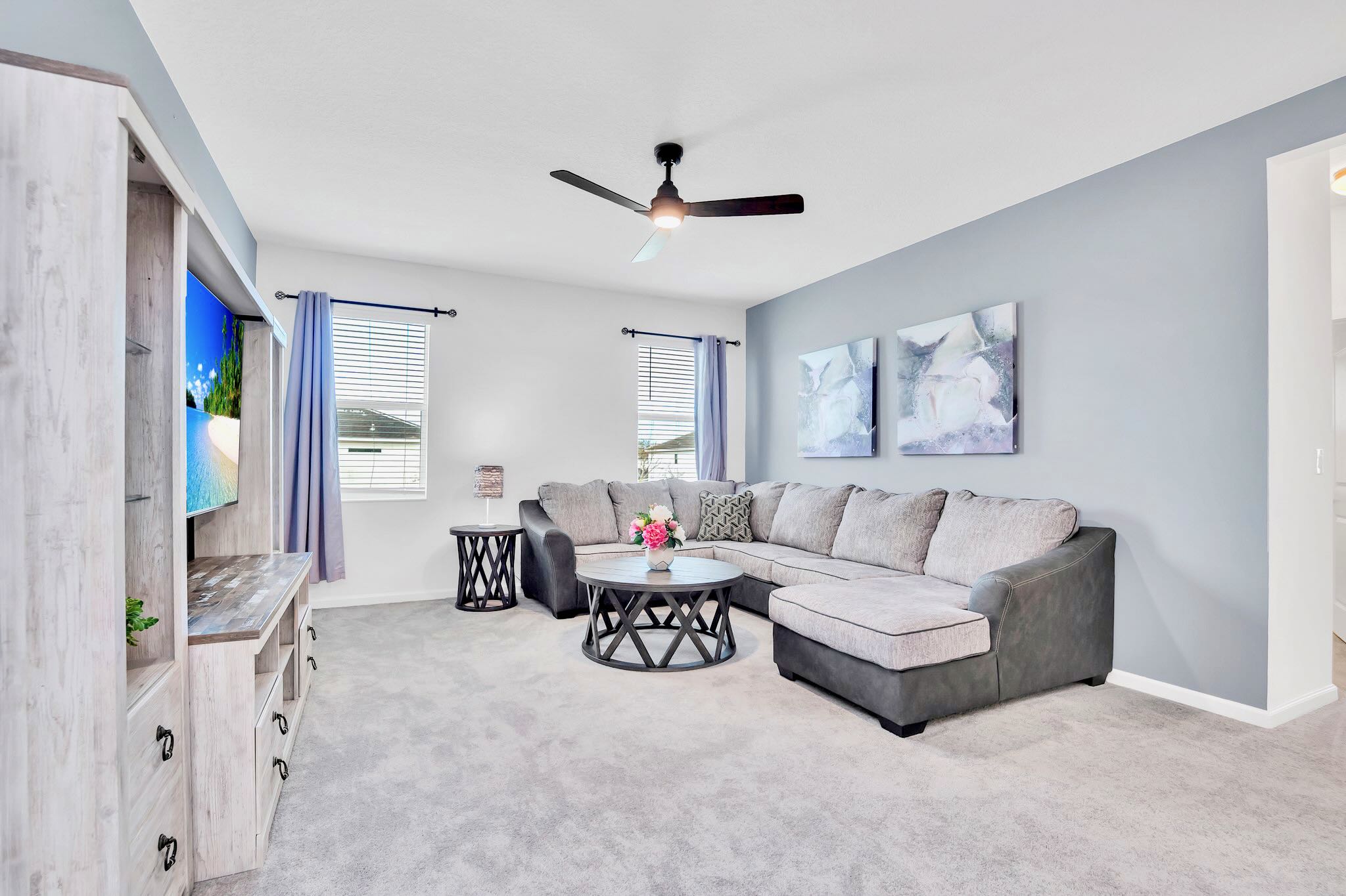 A cozy second living area features a large gray sectional sofa with dark gray trim, centered around a round wooden coffee table. A flat-screen TV is mounted on a rustic entertainment center, all set on light gray carpeted flooring.