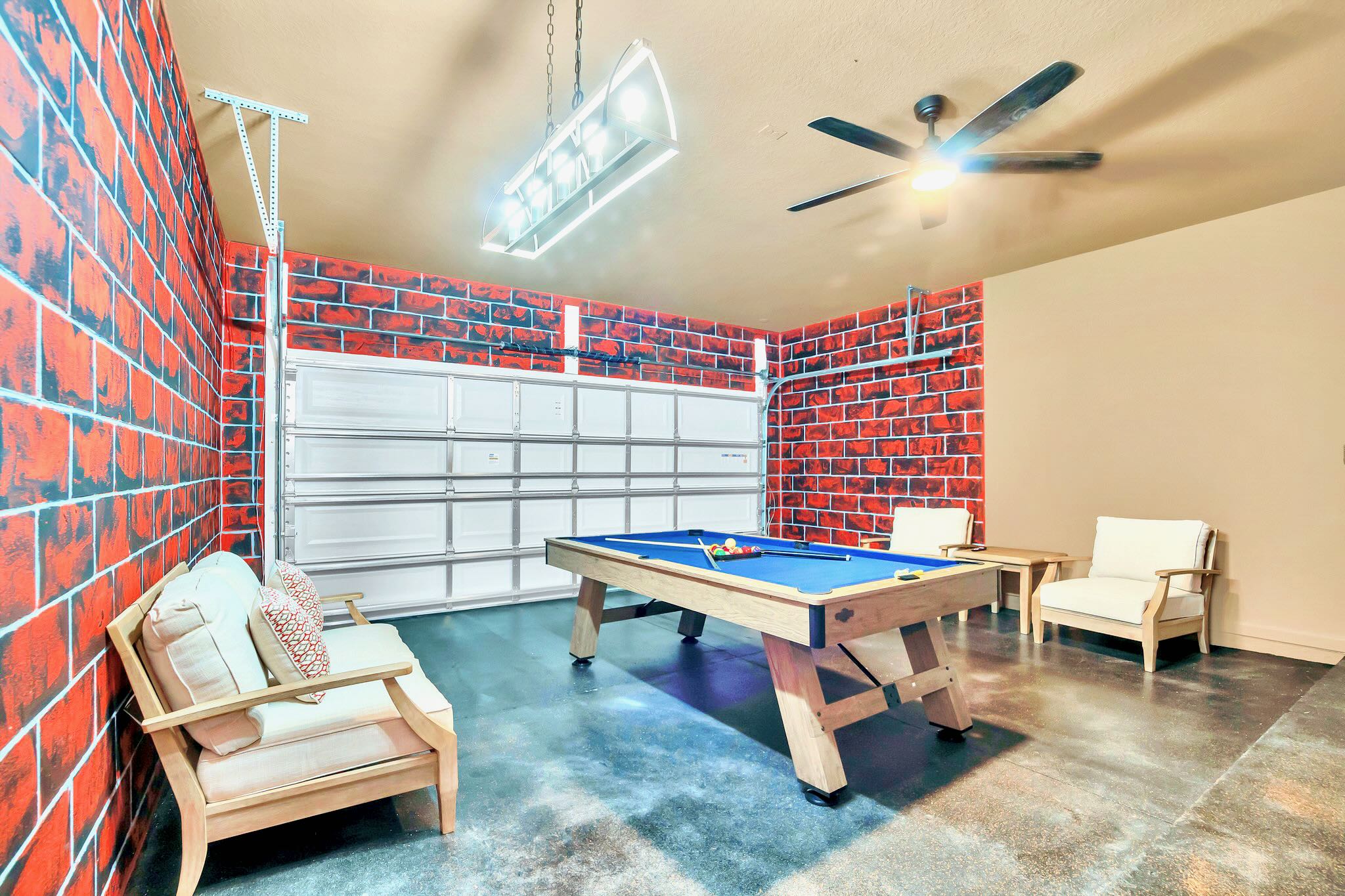 Game room set up in a garage, featuring a pool table with a blue felt surface, exposed brick-patterned walls, and a white garage door. Casual seating and a ceiling fan are also visible.