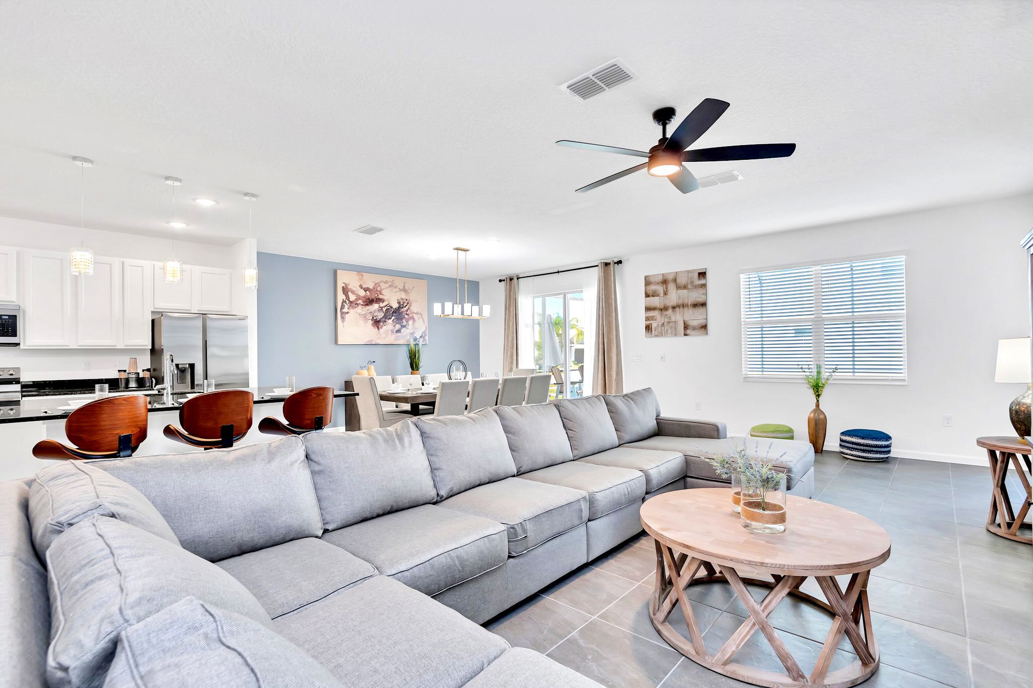 Open-concept living space featuring a large L-shaped gray sectional sofa, a light wood coffee table, and a ceiling fan, with a dining area and kitchen visible in the background. The floor is tiled, and walls are light blue and white.