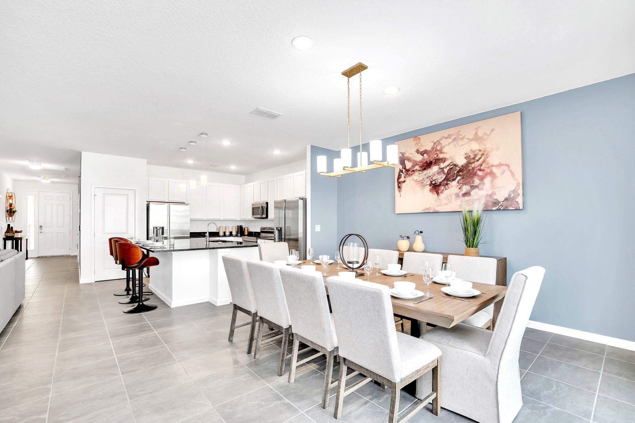 Modern dining area with a long wooden table surrounded by eight upholstered chairs, illuminated by a gold linear chandelier, with a light blue accent wall and abstract art. A white kitchen with a dark island and bar stools is visible to the left.