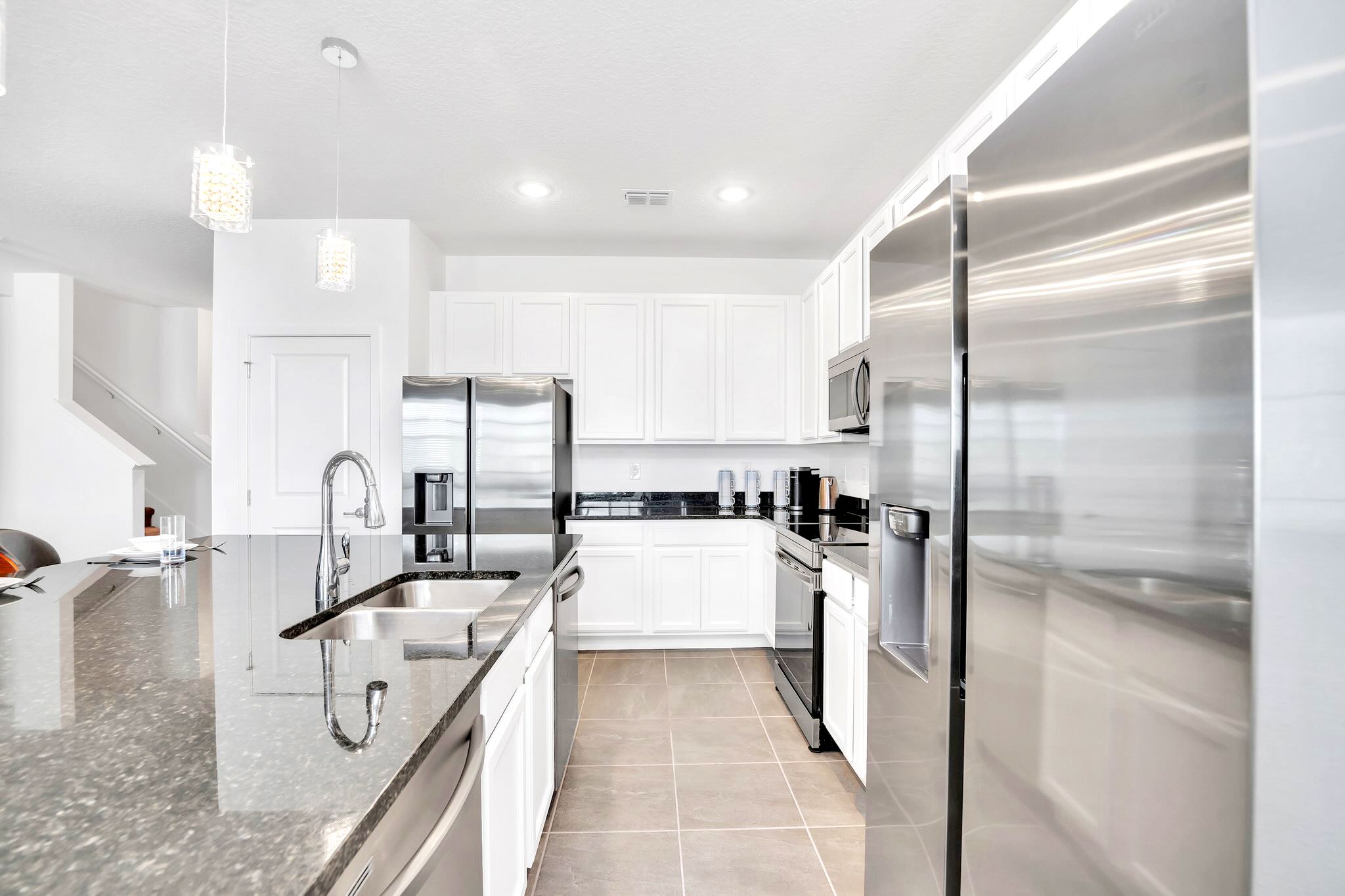 A detailed view of the modern kitchen shows a stainless steel sink set into black granite countertops, surrounded by white shaker cabinets and stainless steel appliances. A dishwasher and a large refrigerator are prominently featured.