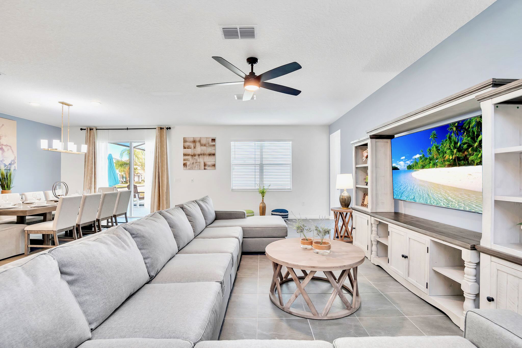 A wide view of the open-concept living space shows a large light gray sectional sofa facing a rustic entertainment center with a TV, and a round wooden coffee table. Sliding glass doors lead to an outdoor area, with a dining table visible to the left.