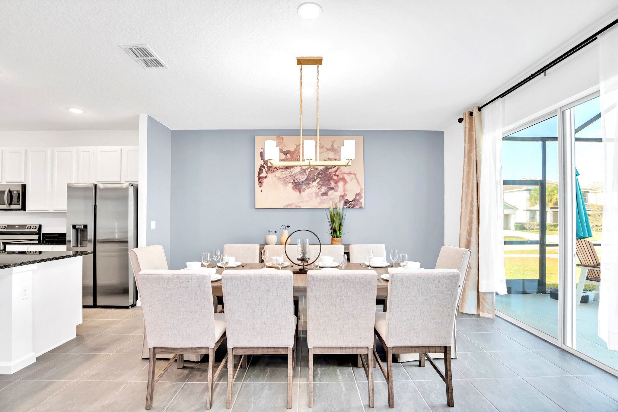 A modern dining area features a long rectangular wooden table set for eight with light gray upholstered chairs, illuminated by a gold linear chandelier. A large abstract artwork hangs on the light blue accent wall, and sliding glass doors open to an outdoor space.