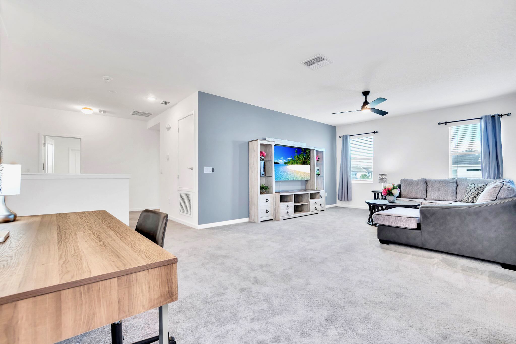 An expansive view of the second living room highlights a large gray sectional sofa facing a TV on a rustic entertainment center, with light blue accent walls. A wooden desk and chair are visible to the left, all on light gray carpeted flooring.